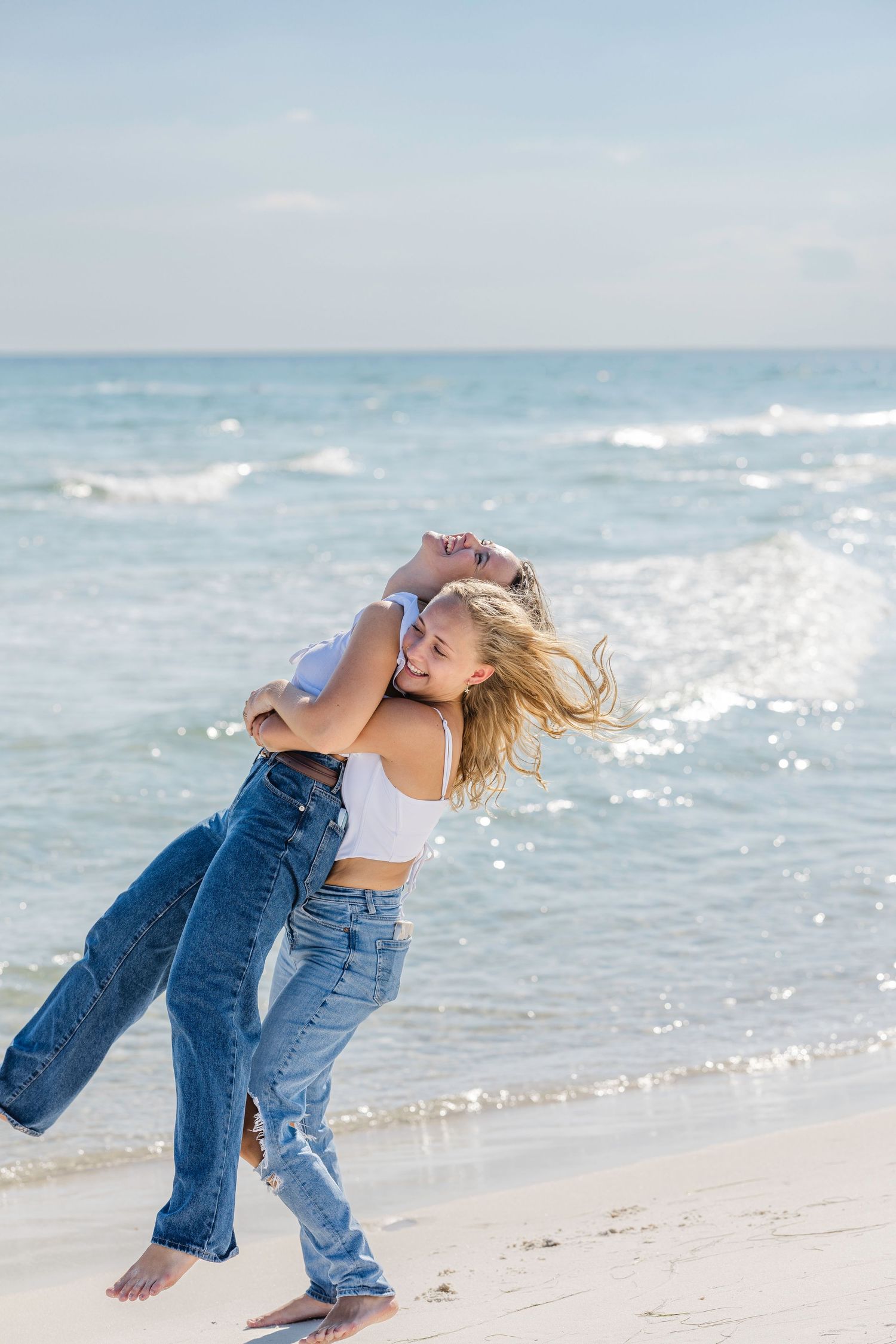 Dancing figure in denim and white crop top twirls joyfully on a sunny beach with ocean waves in the background.