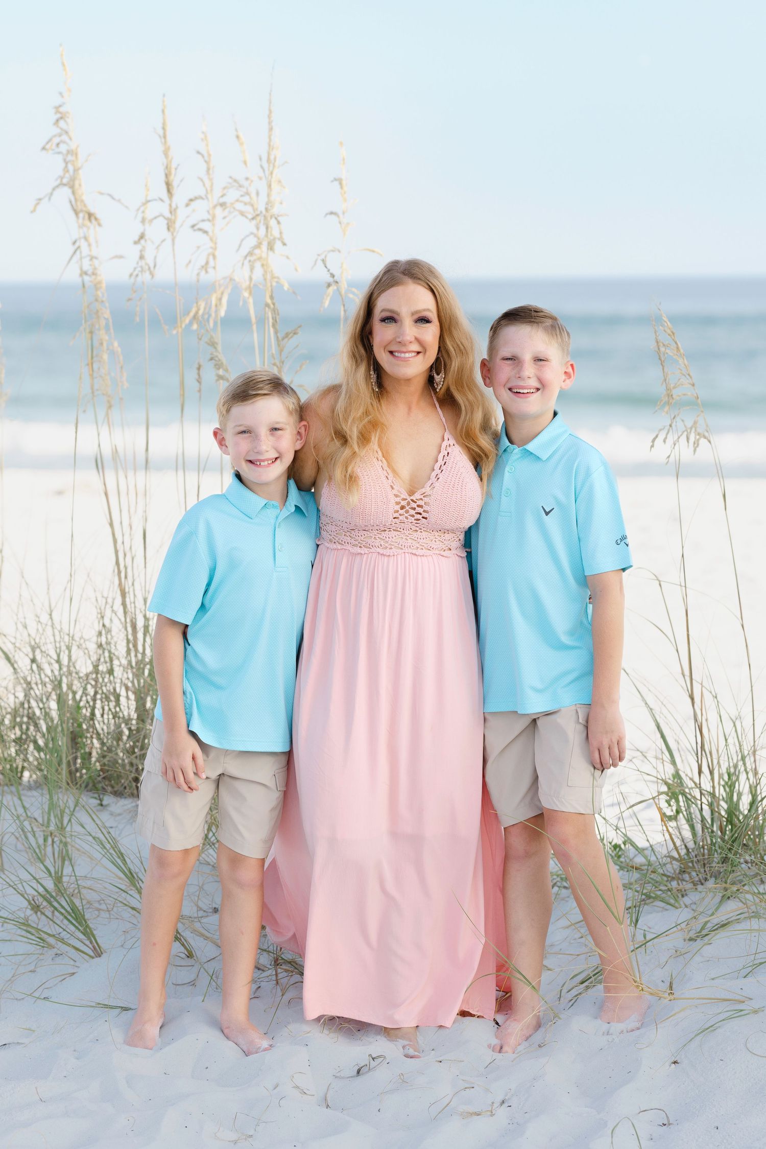 A beachside family portrait with two children in blue polos and a woman in a pink maxi dress among sea oats.