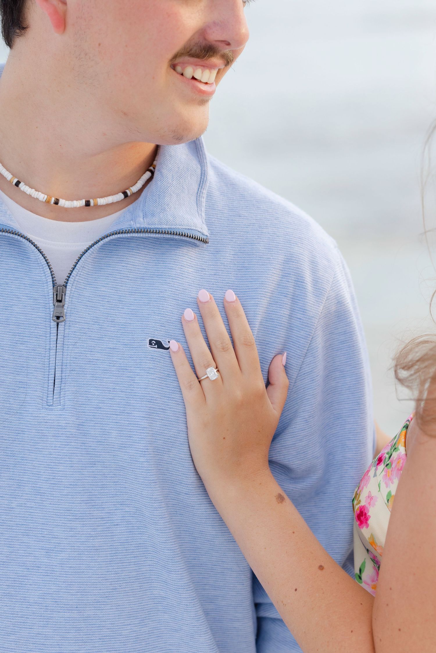 Close up view of an engagement ring on a hand resting against a light blue shirt by the beach.