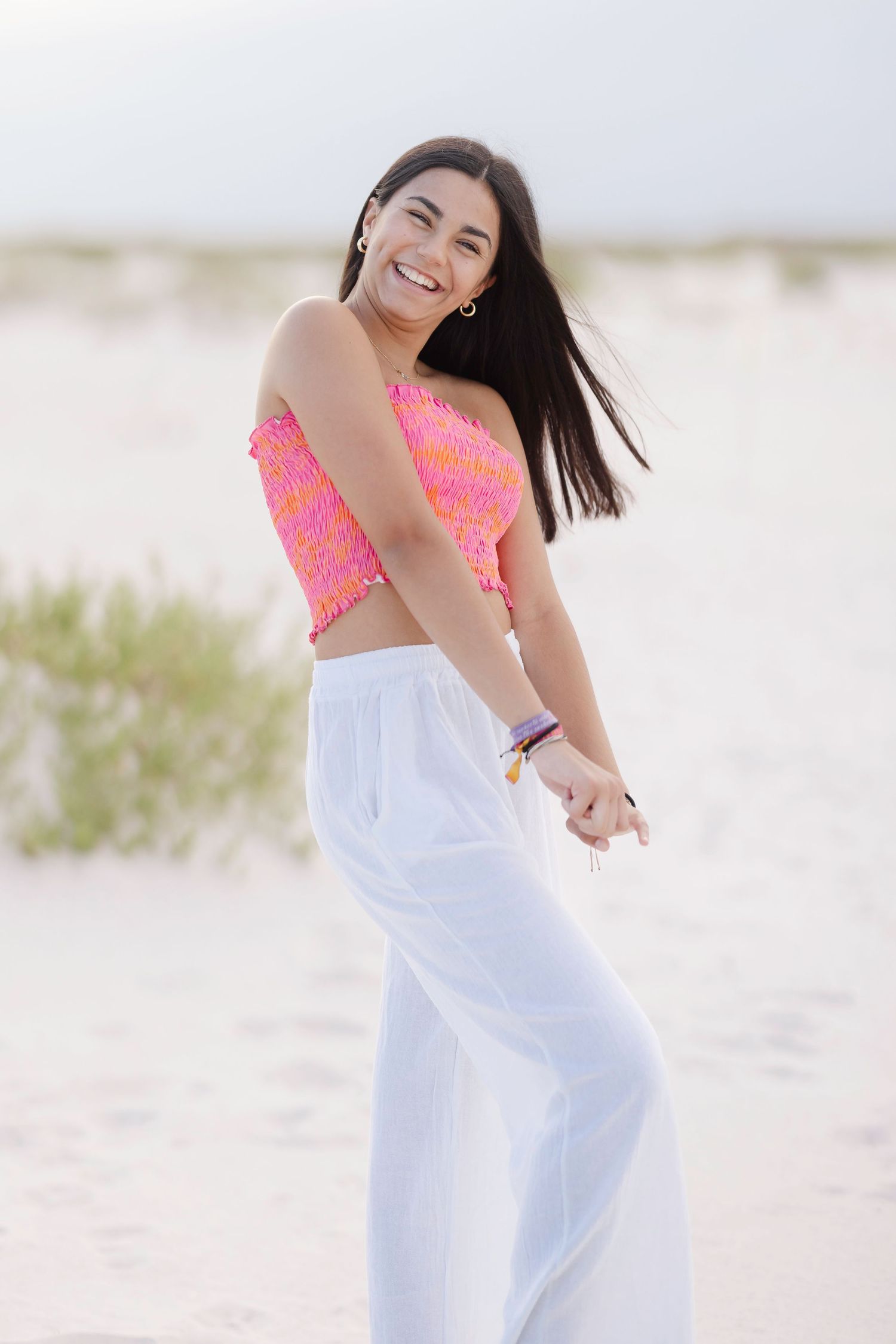 Someone wearing a coral crop top and white flowy pants poses joyfully on a beach with windswept hair.