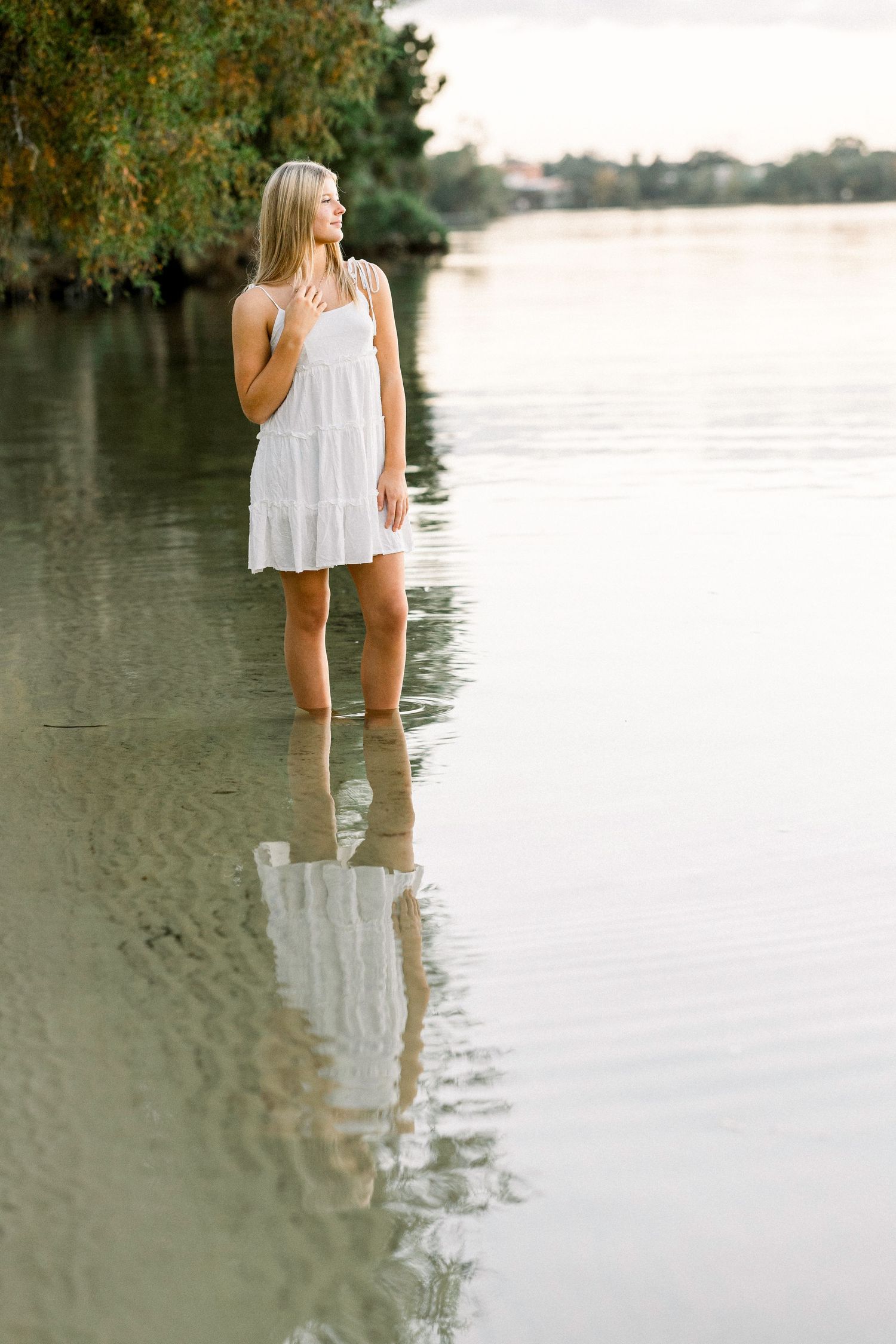 Figure in a white summer dress stands ankle-deep in calm lake water at sunset, creating a peaceful reflection.