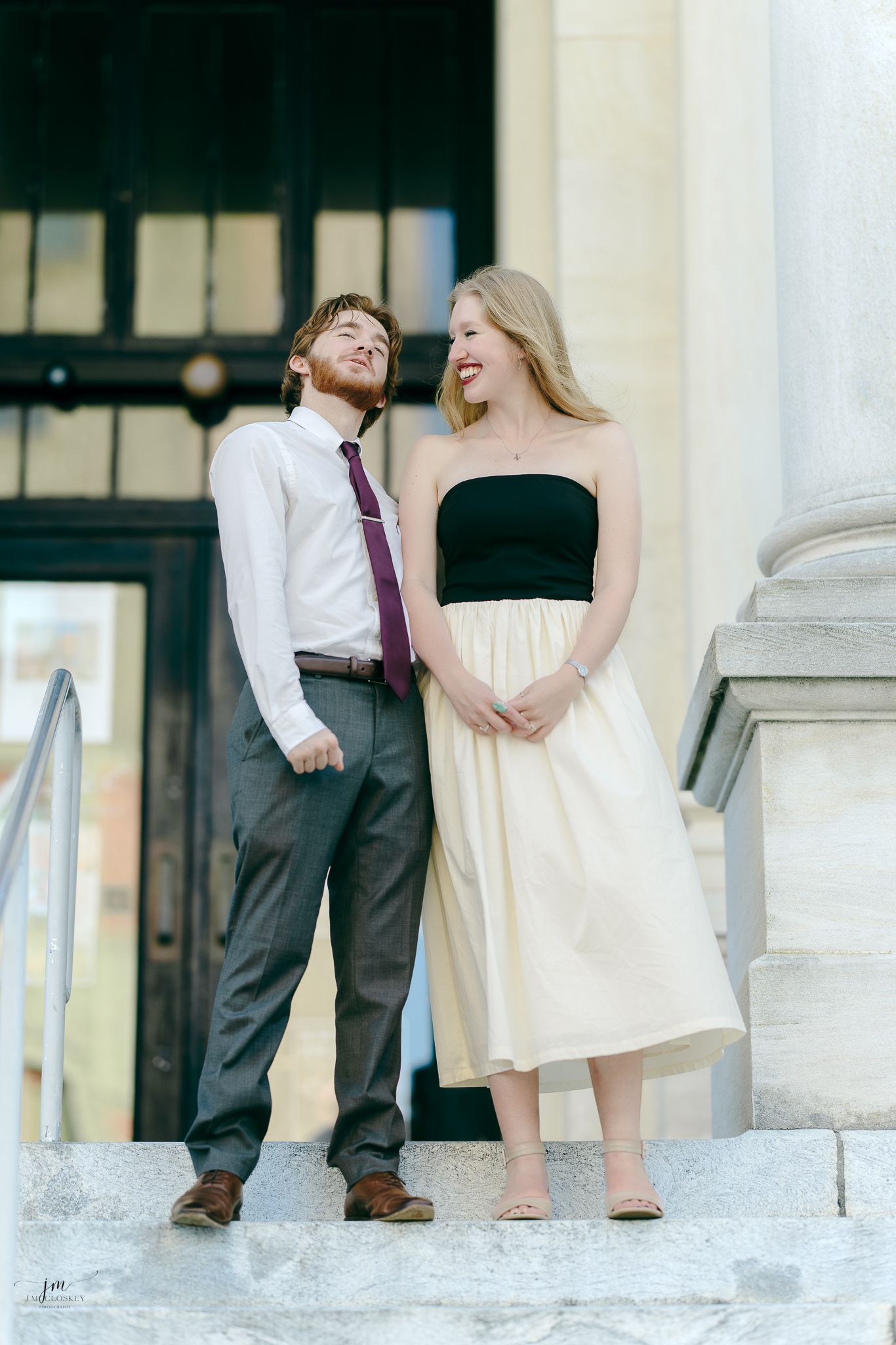 A couple in formal attire shares a lighthearted moment while standing on stone steps outside a building.