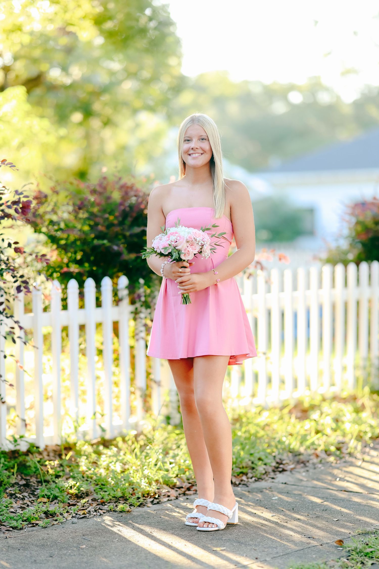 Someone in a pink summer dress holding a bouquet stands next to a white picket fence on a sunny day.