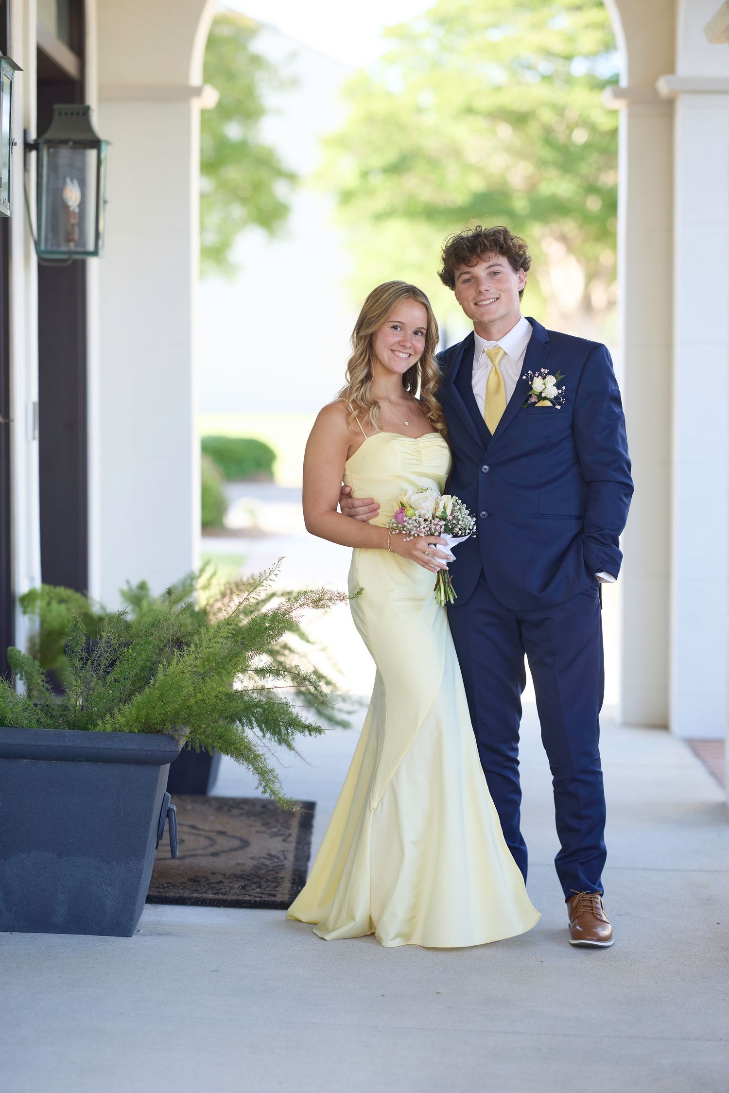 A couple poses together at a dance event, with one wearing a pale yellow dress and one in a navy suit and tie.