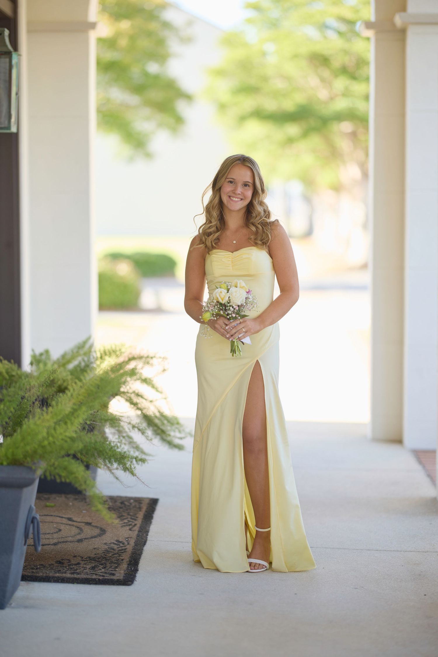 Bridesmaids in flowing pale yellow dresses carry bouquets while walking through a covered outdoor colonnade.