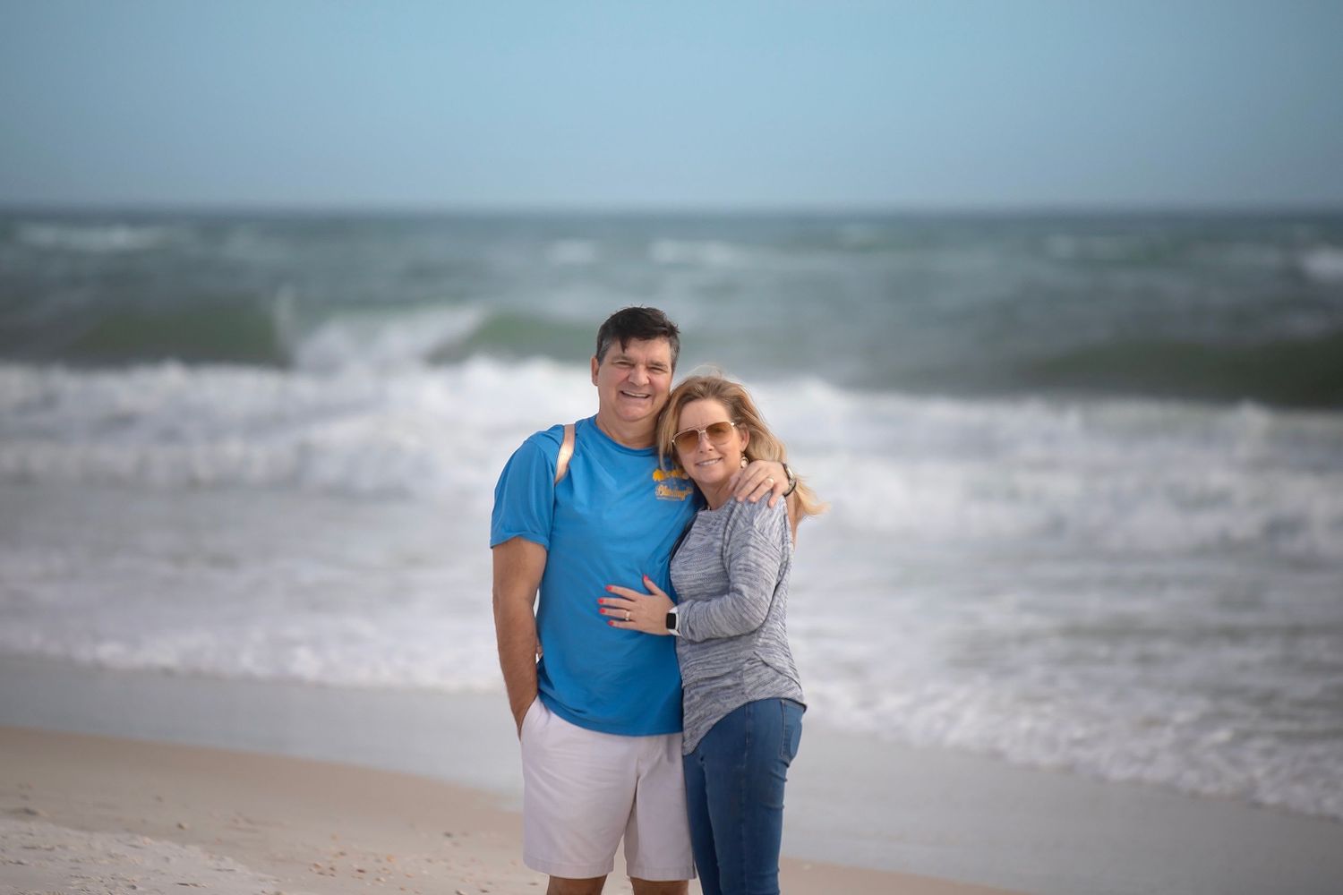 A couple embraces on a beach with crashing waves and overcast skies in the background.
