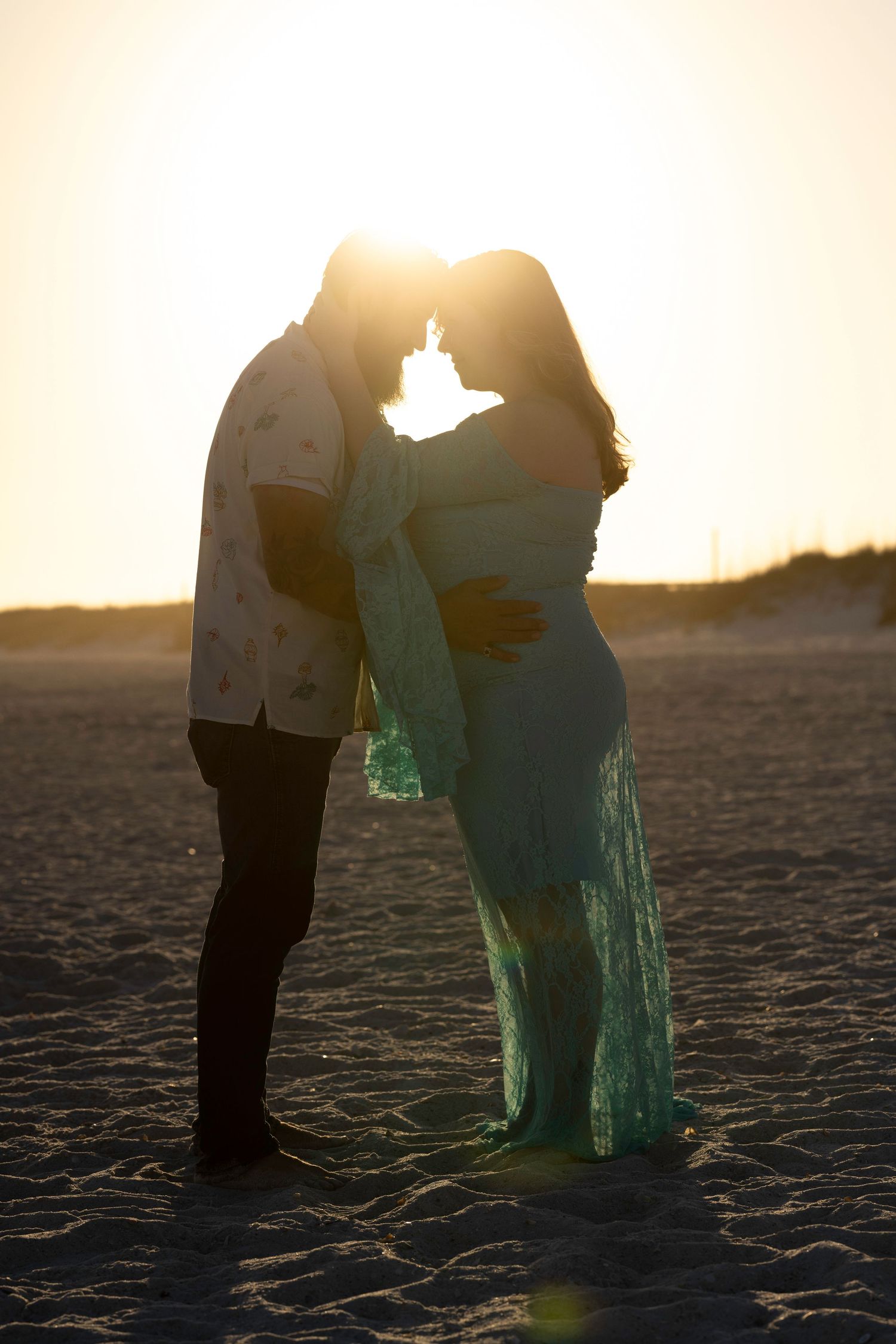 Couple sharing a romantic sunset moment on the beach, woman in flowing teal maternity gown, backlit by golden light.