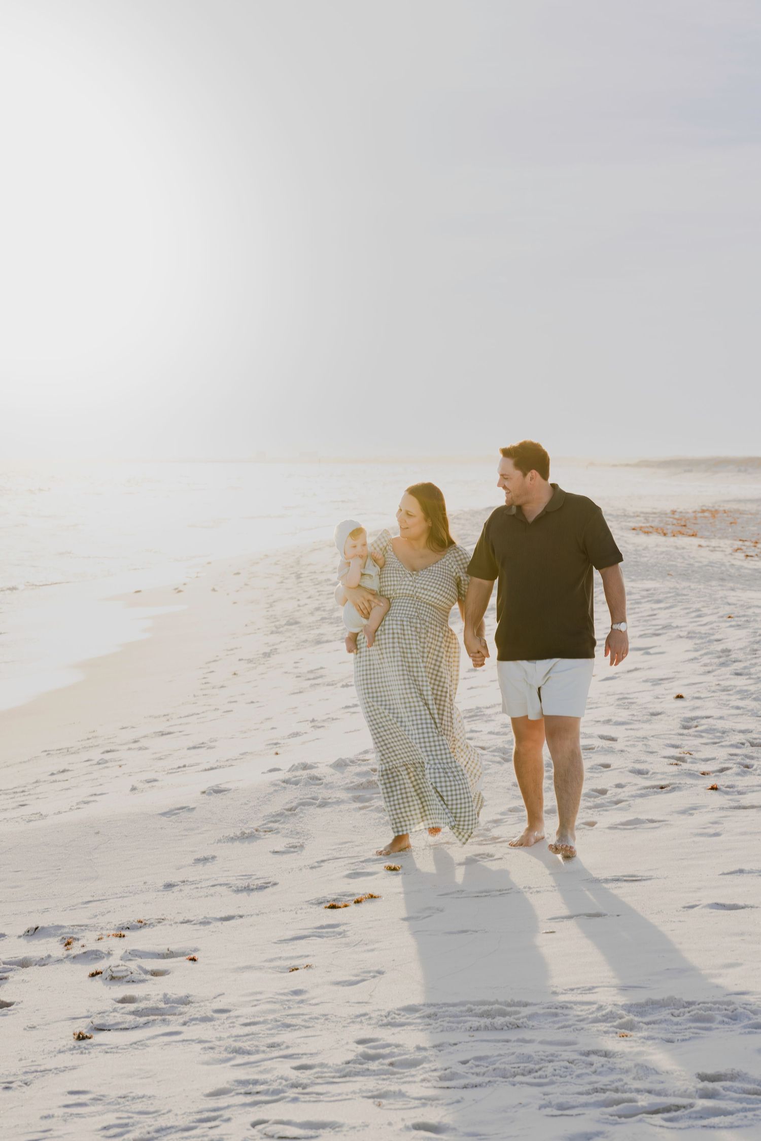 A young family walks barefoot on a sunny white sand beach, mother holding a baby while father strolls beside them.