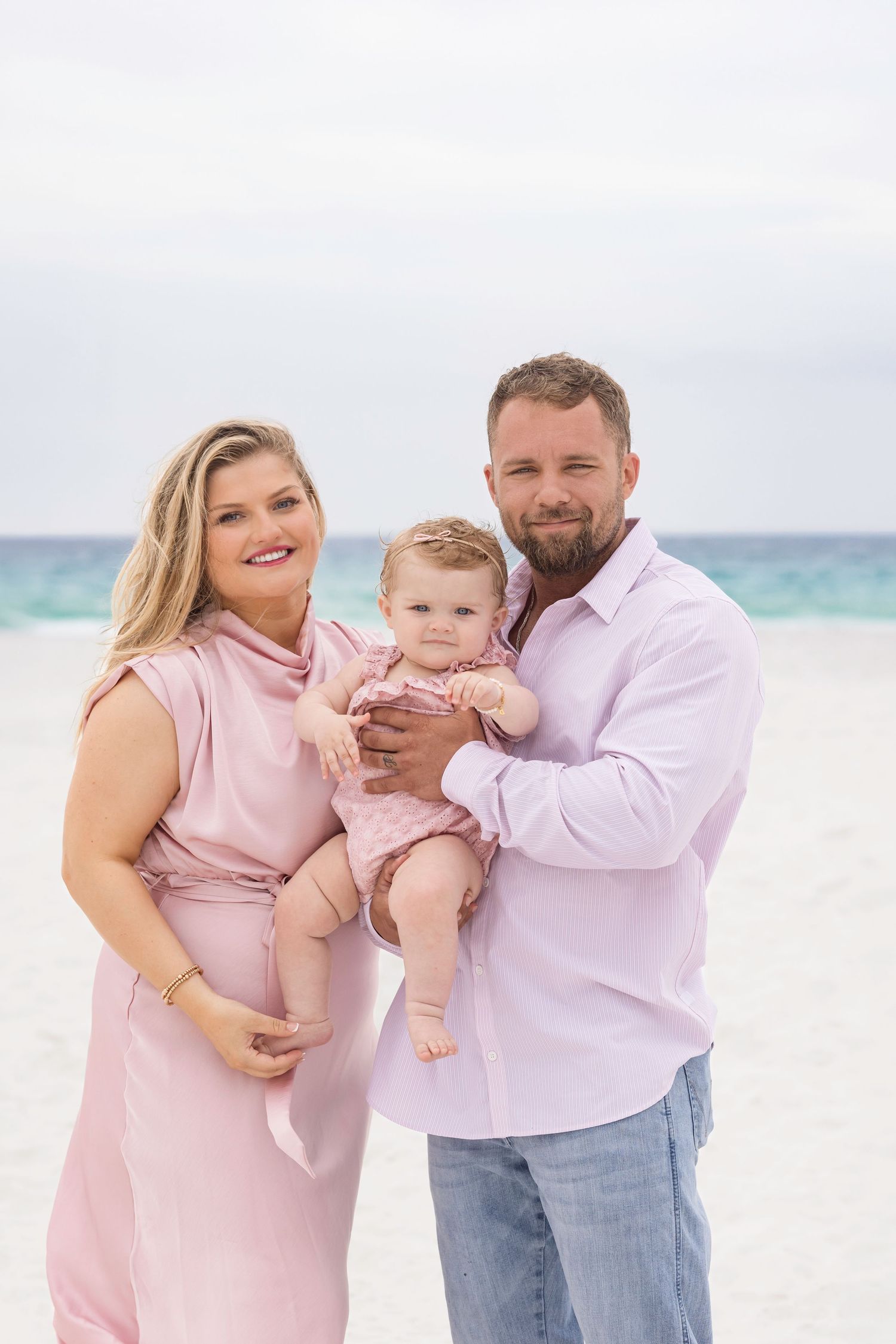 A happy family of three poses on a white sand beach, with parents in pink outfits holding their baby girl.