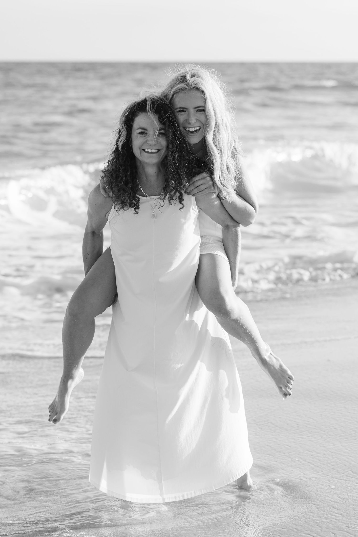 Two women laughing on beach, one giving piggyback ride, wearing white dress, ocean waves behind them, black and white photo.
