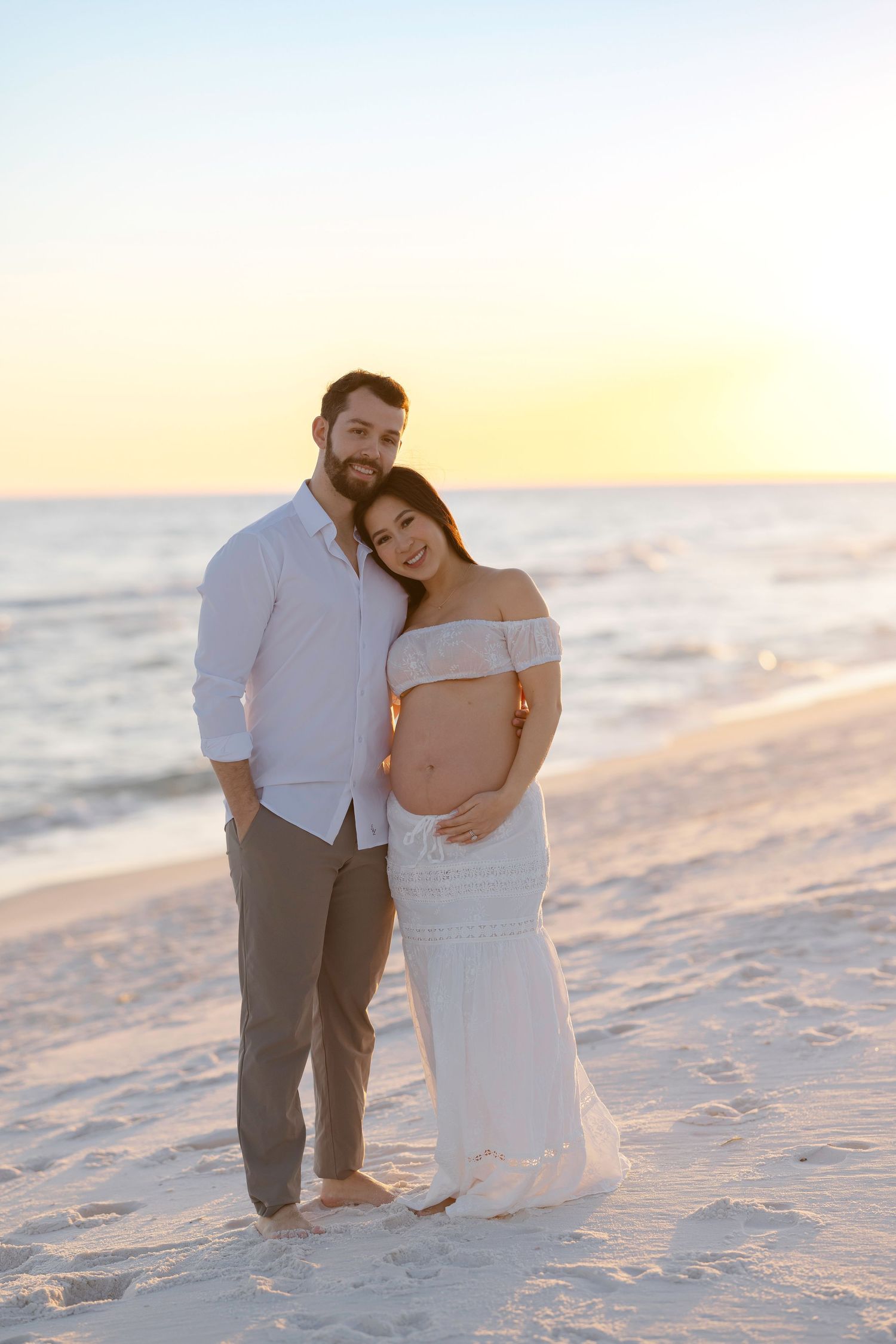 A romantic couple in white and gray attire embraces on a sandy beach at sunset.