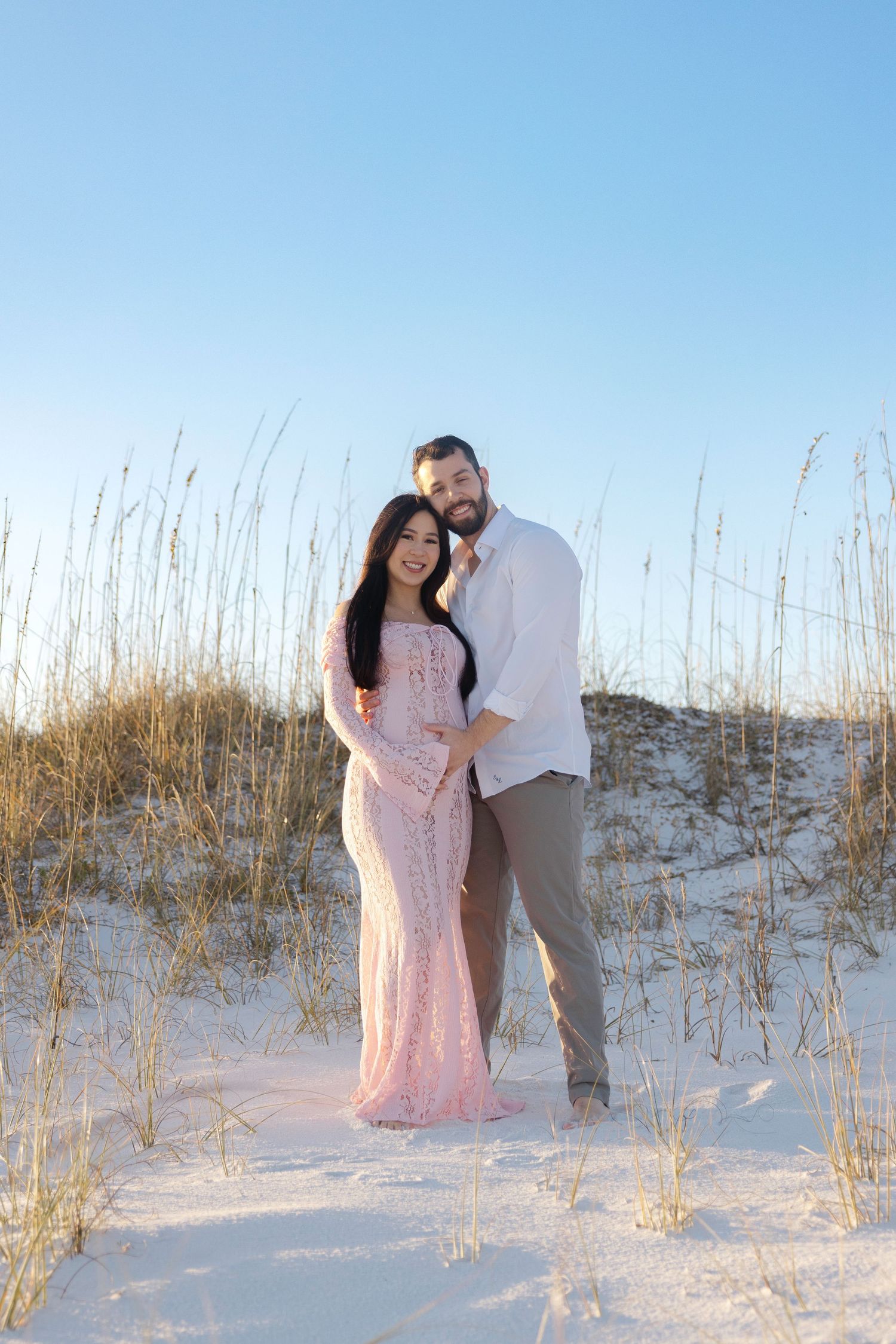 Couple posing together among beach dunes with tall grass during golden hour at the seashore.
