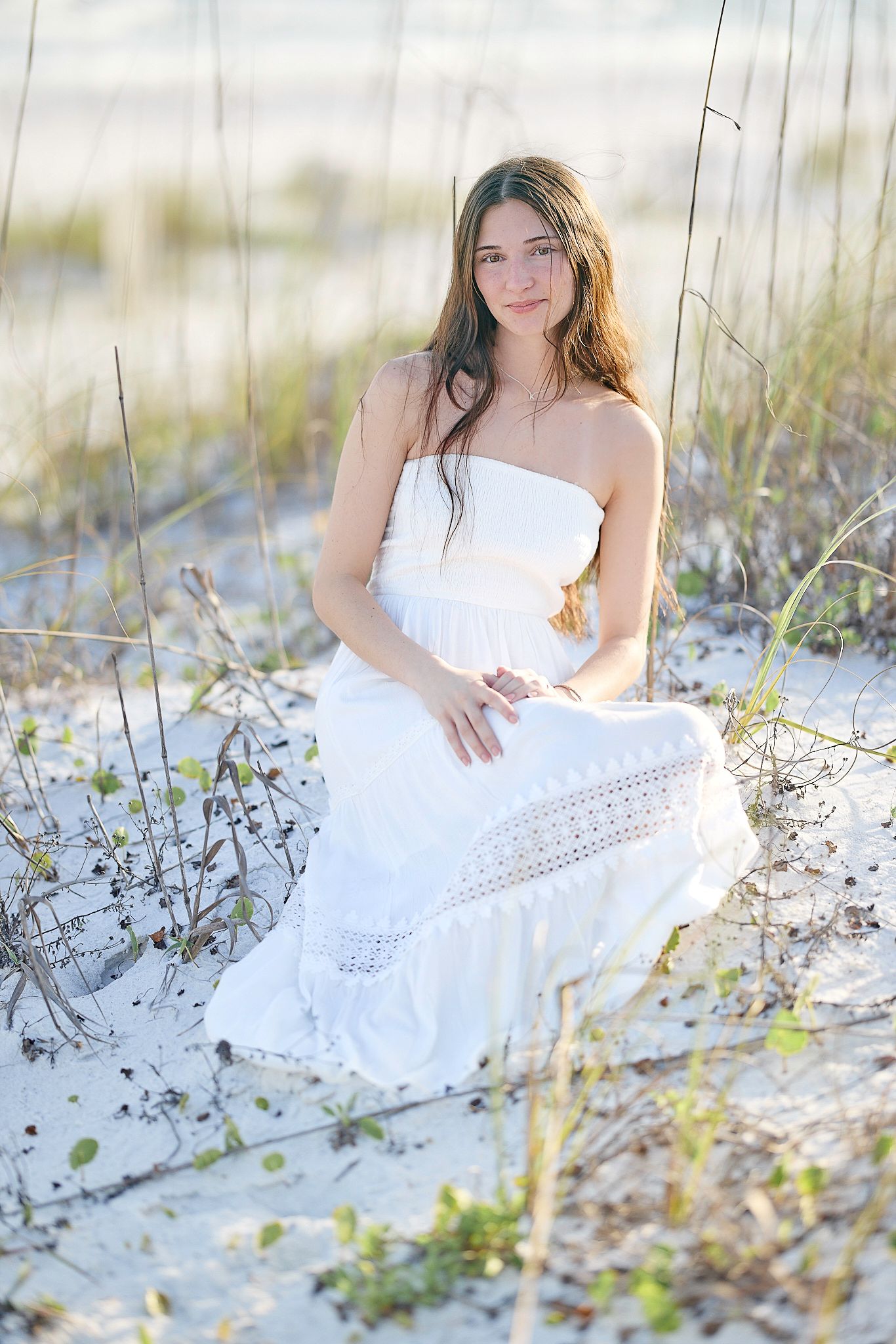 A white strapless sundress is worn while sitting in soft sand among beach grass on a sunny summer day.