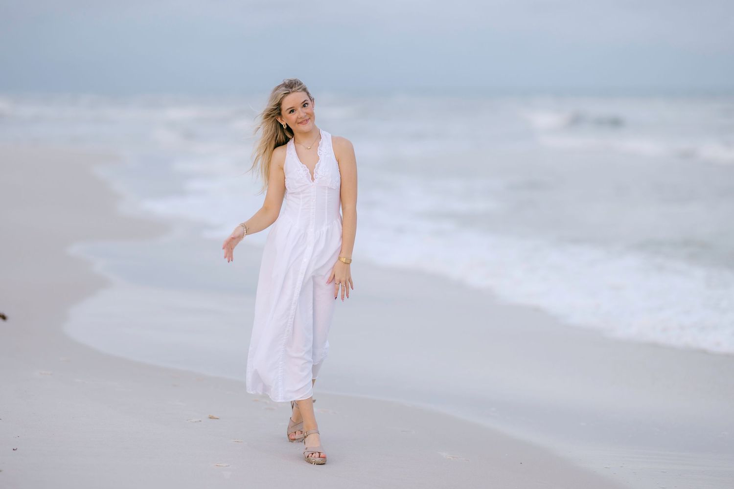 Someone in a flowing white dress walks along a serene beach at dusk with waves rolling in the background.