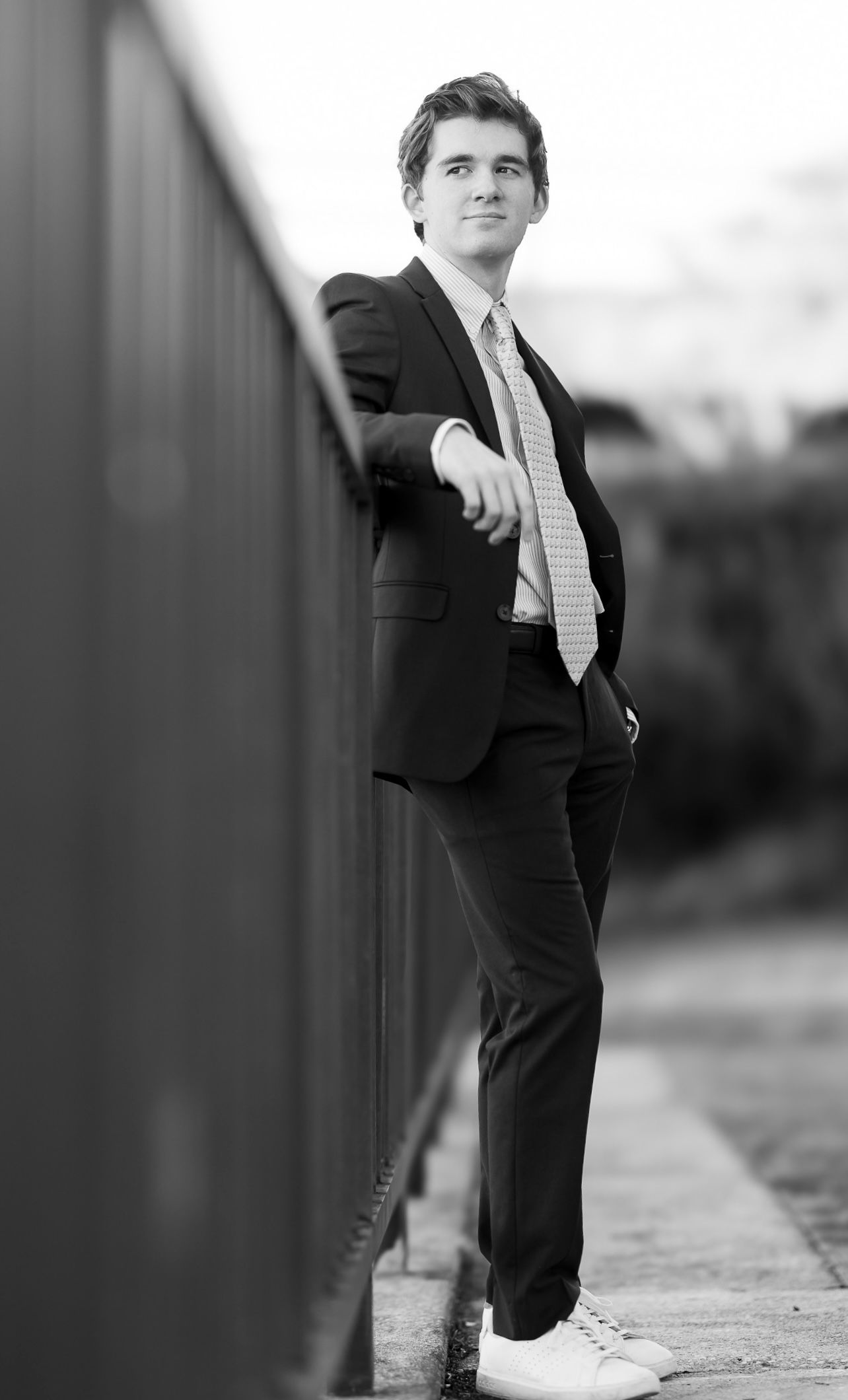 Black and white photo of a businessman in a dark suit and tie leaning casually against a railing outdoors.