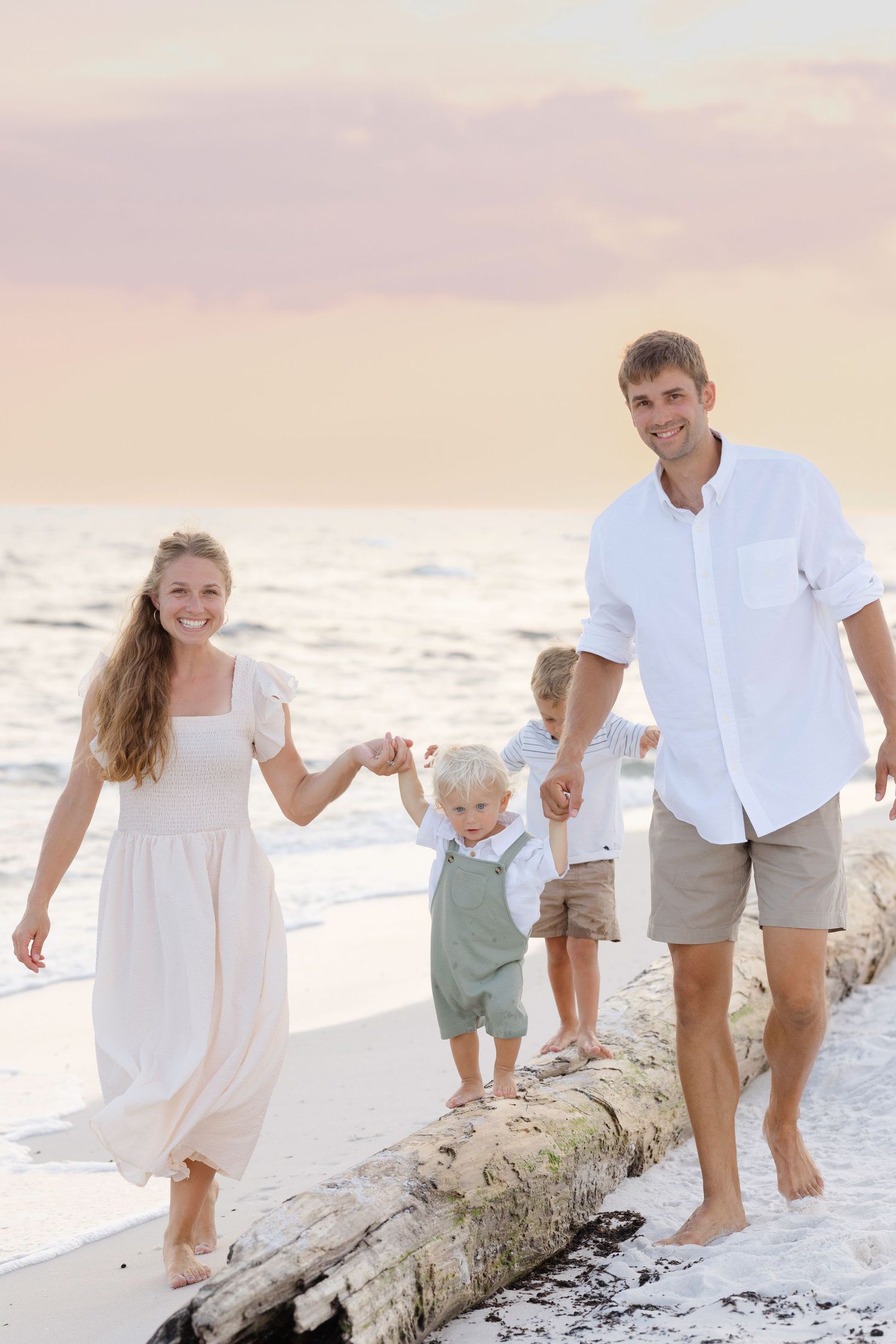 Family playing together on a driftwood log at the beach during sunset wearing coordinated white outfits.