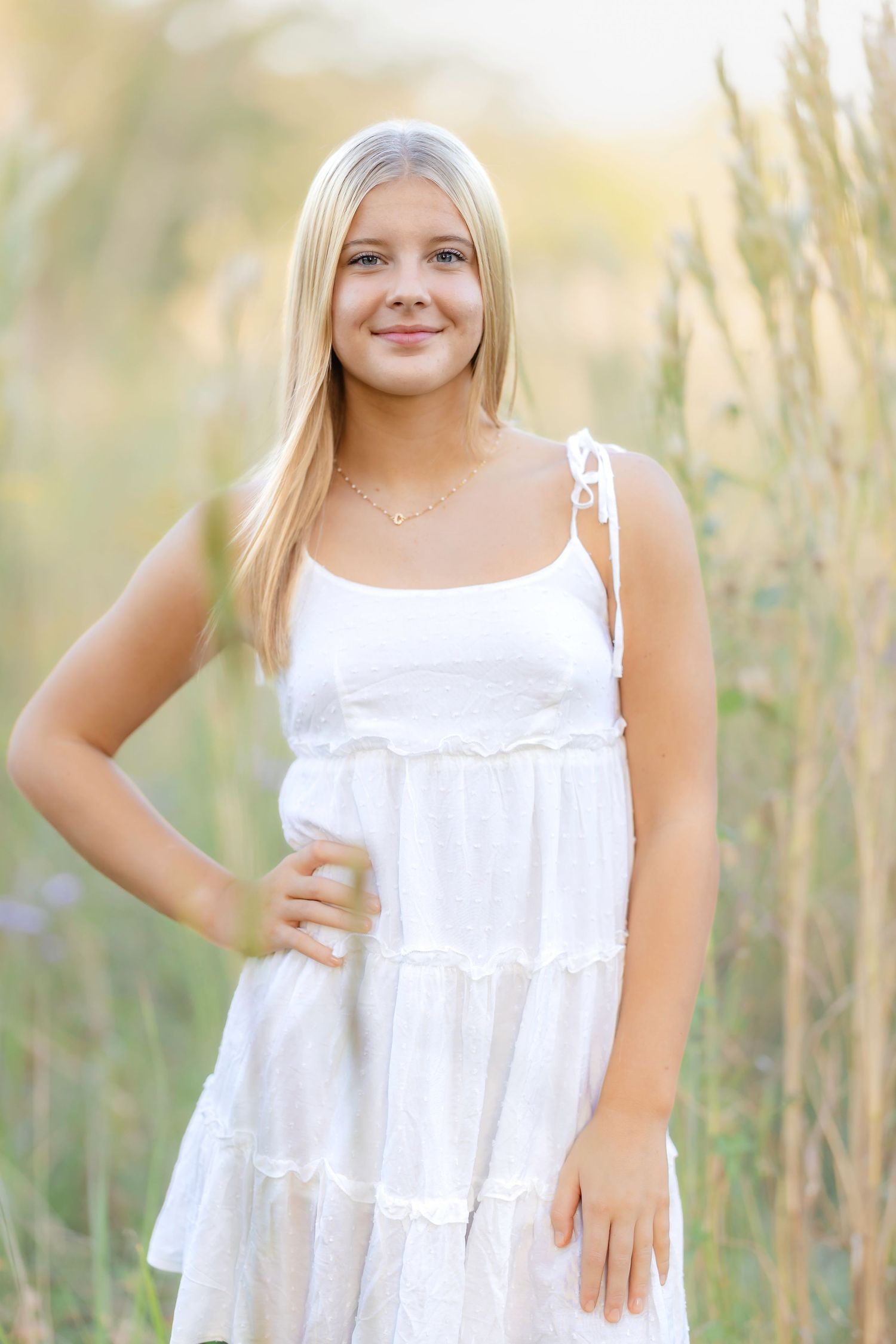 Smiling blonde teen in white sundress standing in tall grass field in warm sunlight.