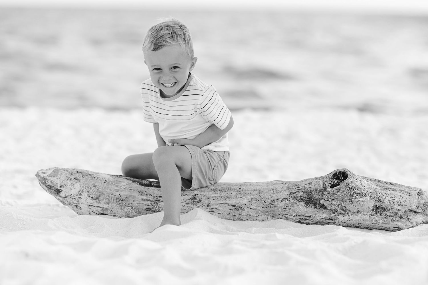 Black and white photo of a smiling boy sitting on driftwood on a sandy beach.