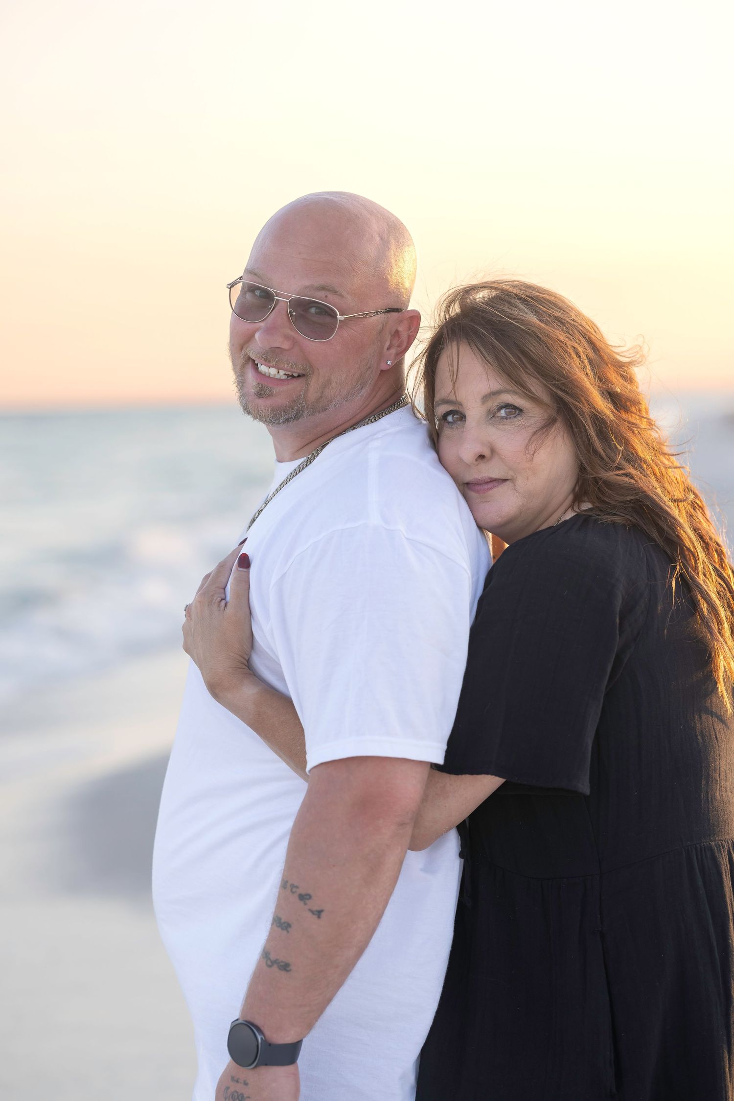 A bald man with sunglasses and a woman in black embrace on a beach at golden sunset.