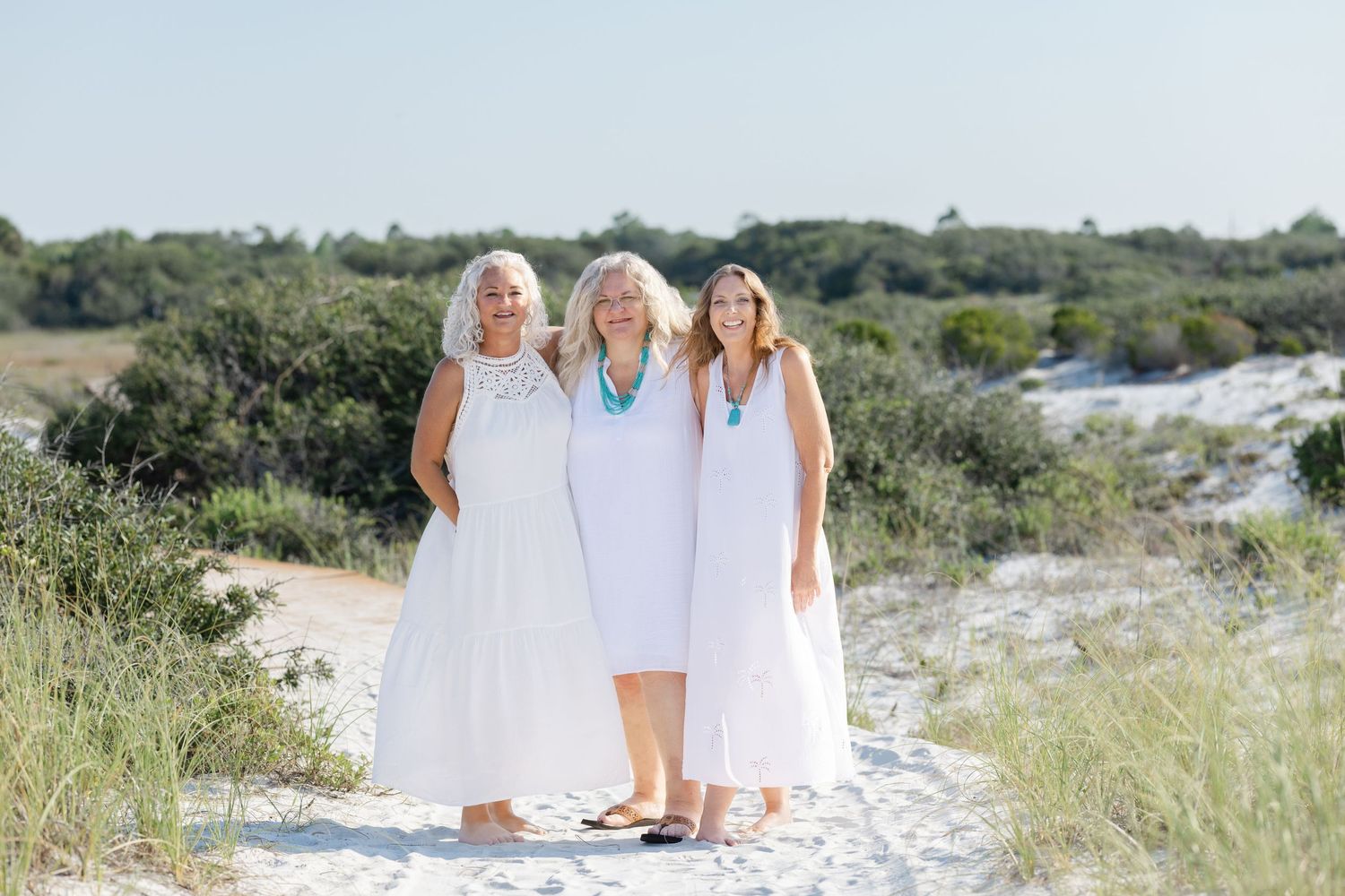Three people in white dresses stand together on a sandy beach path surrounded by coastal vegetation.