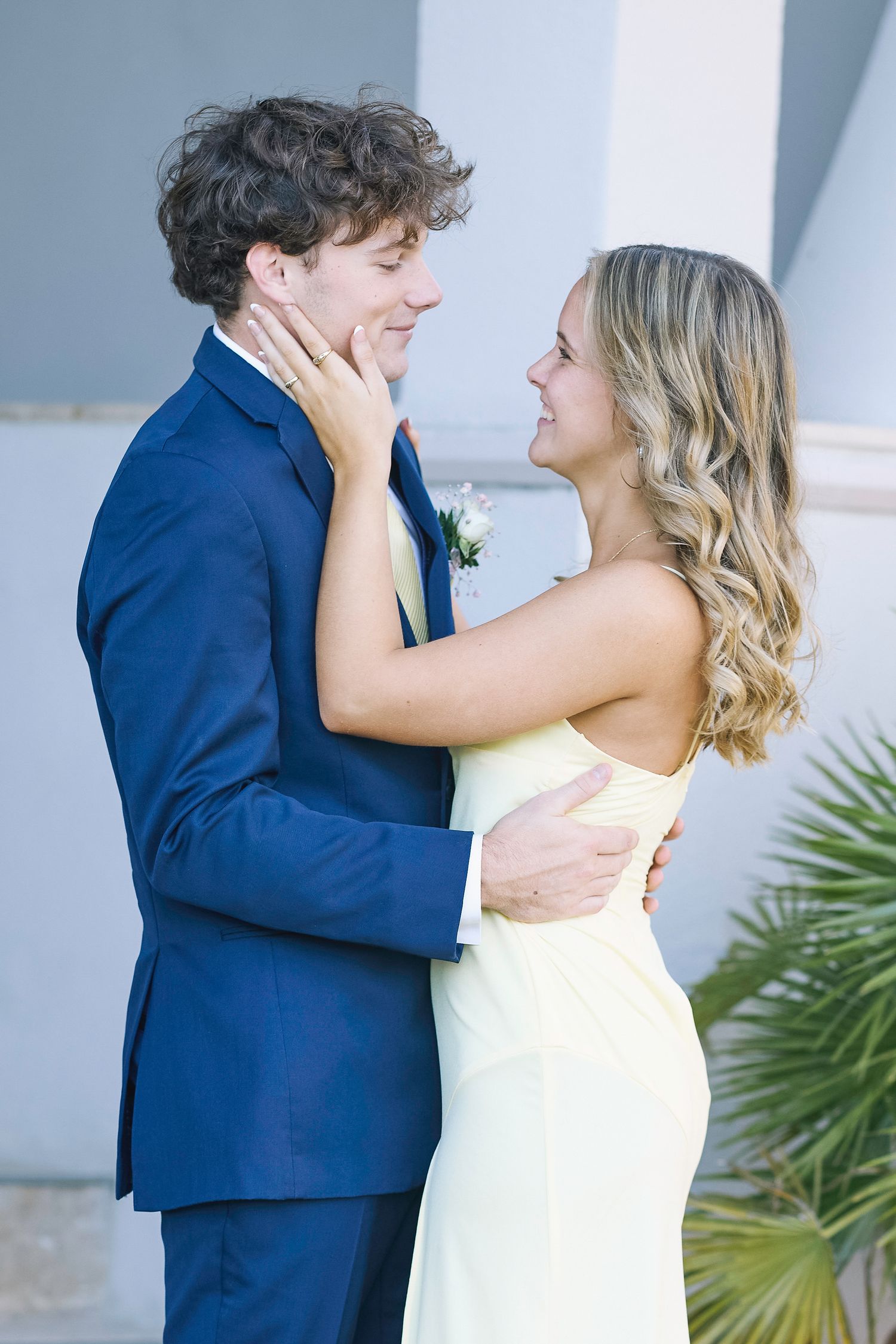 A couple in navy blue suit and cream dress embrace tenderly against a light colored wall with tropical foliage.