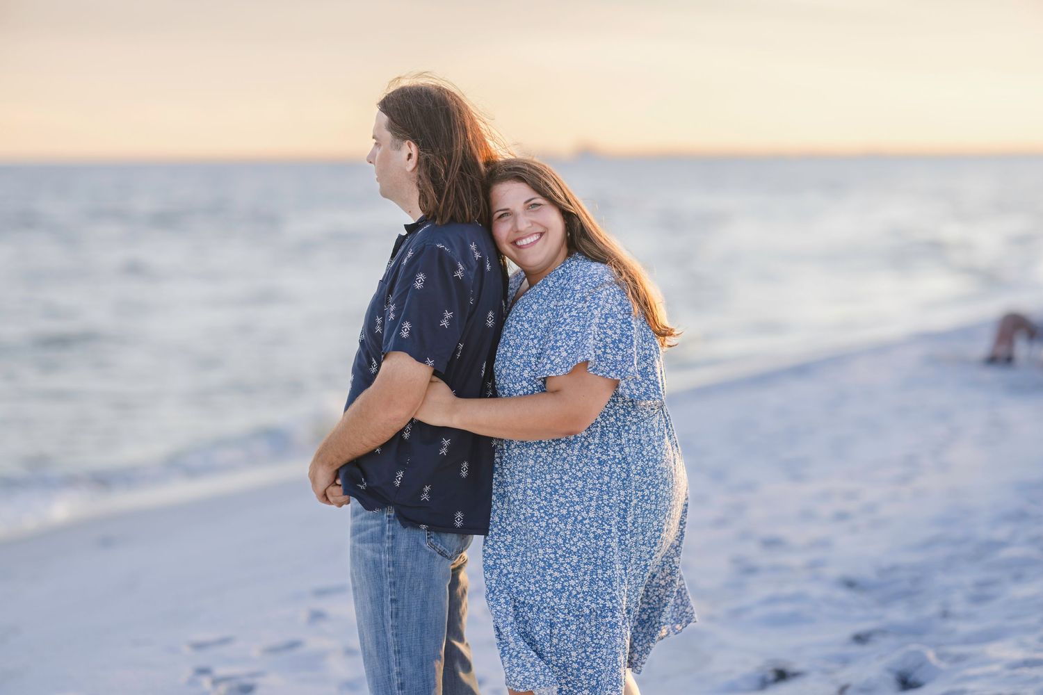 A couple embraces and smiles on a sandy beach at sunset, the woman in a blue floral dress hugging the man from behind.