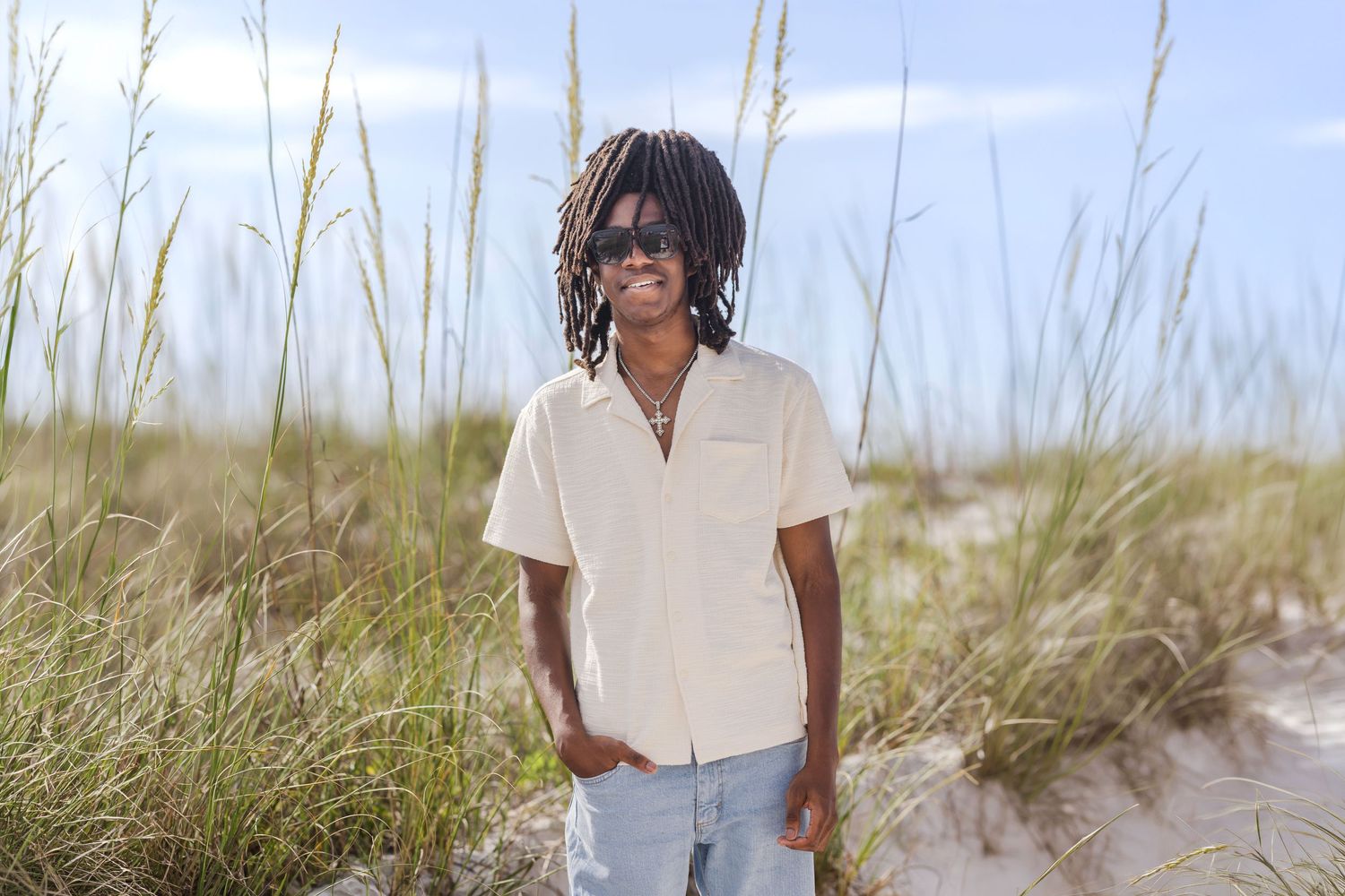 Young man with dreadlocks and sunglasses smiling on a sunny beach with tall grass and blue sky.