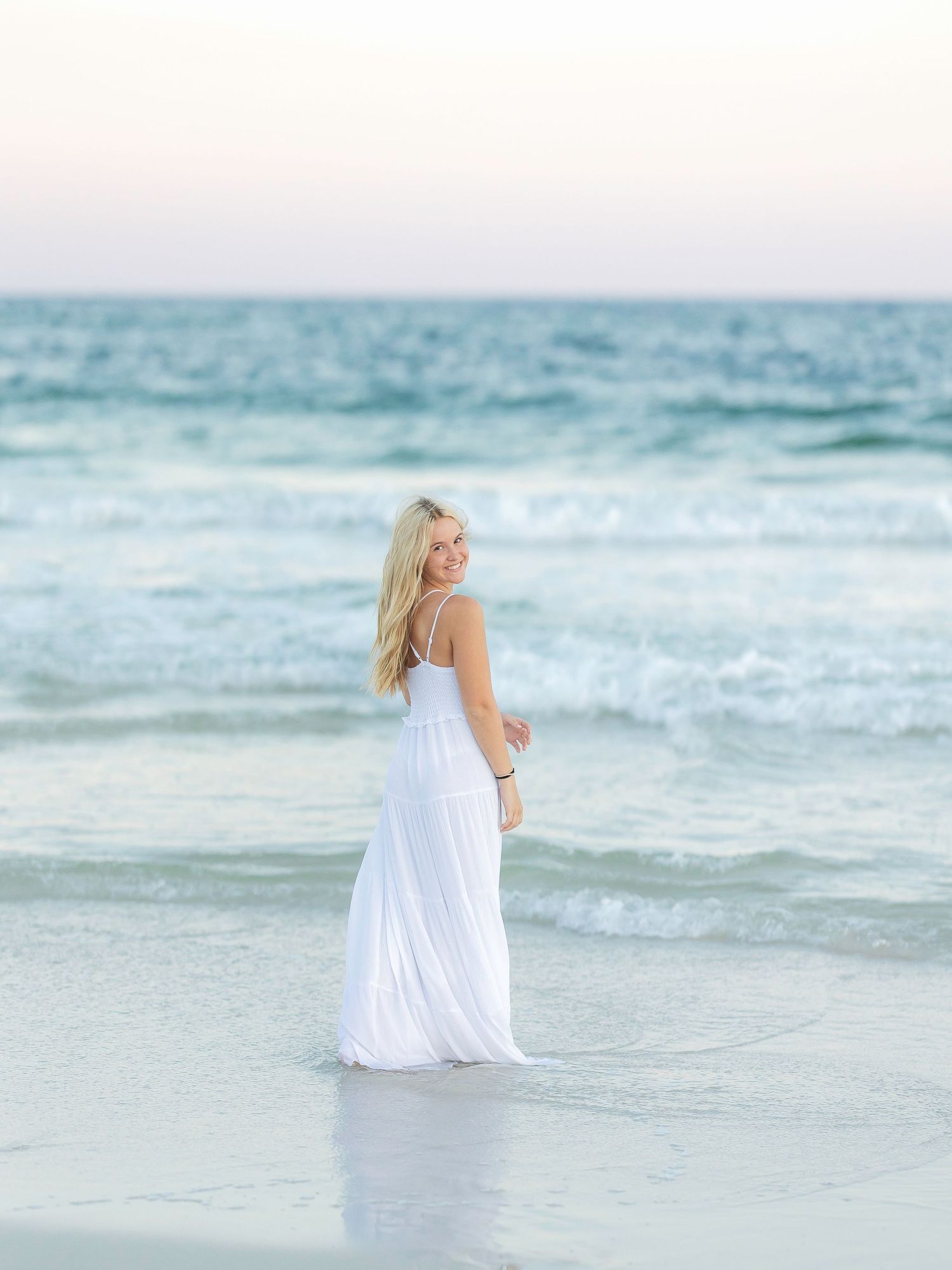 Blonde woman in white maxi dress standing in ocean waves on a serene beach at sunset.