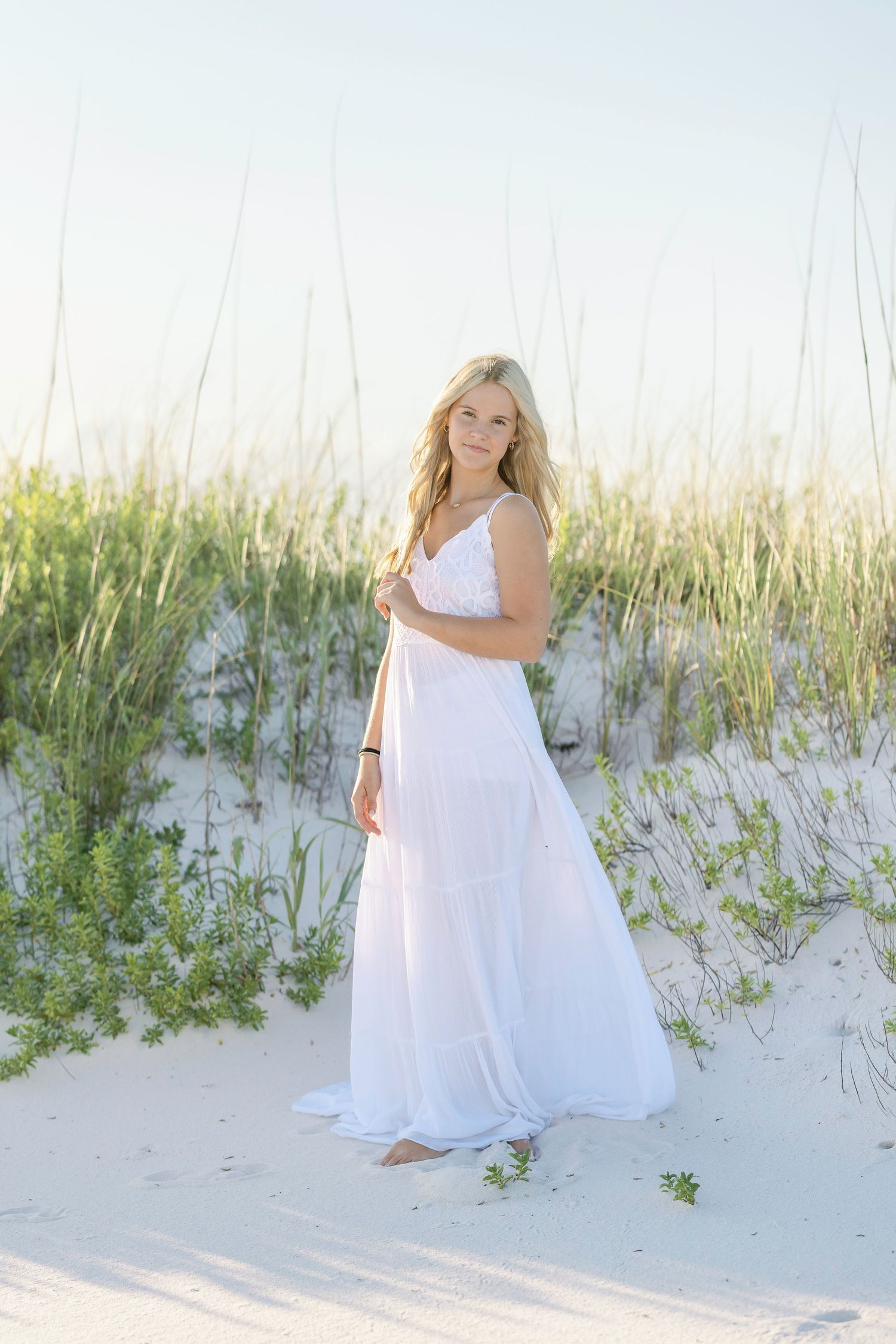 Blonde woman in white maxi dress standing on sandy beach dunes with sea grass.