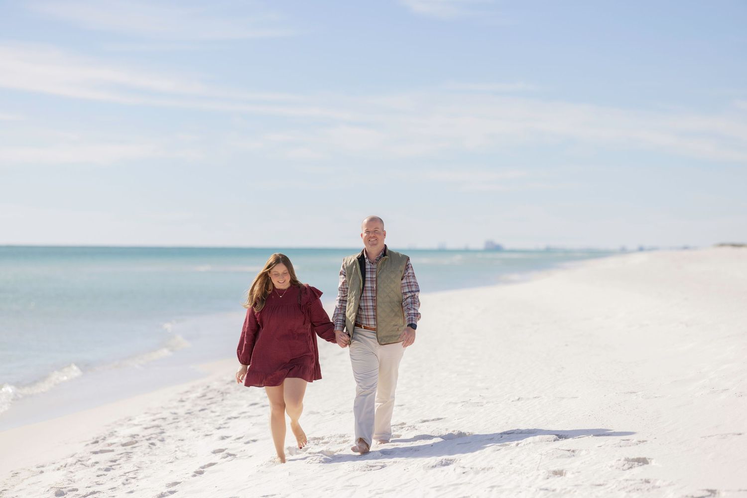 Couple holding hands walking on a white sand beach with calm blue ocean waters on a clear sunny day.