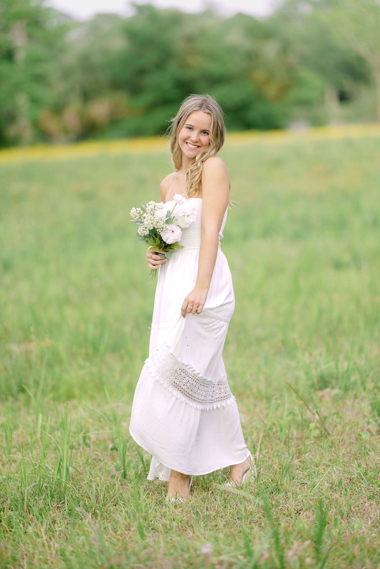 Smiling blonde woman in white strapless maxi dress holding flower bouquet while walking barefoot through green meadow.
