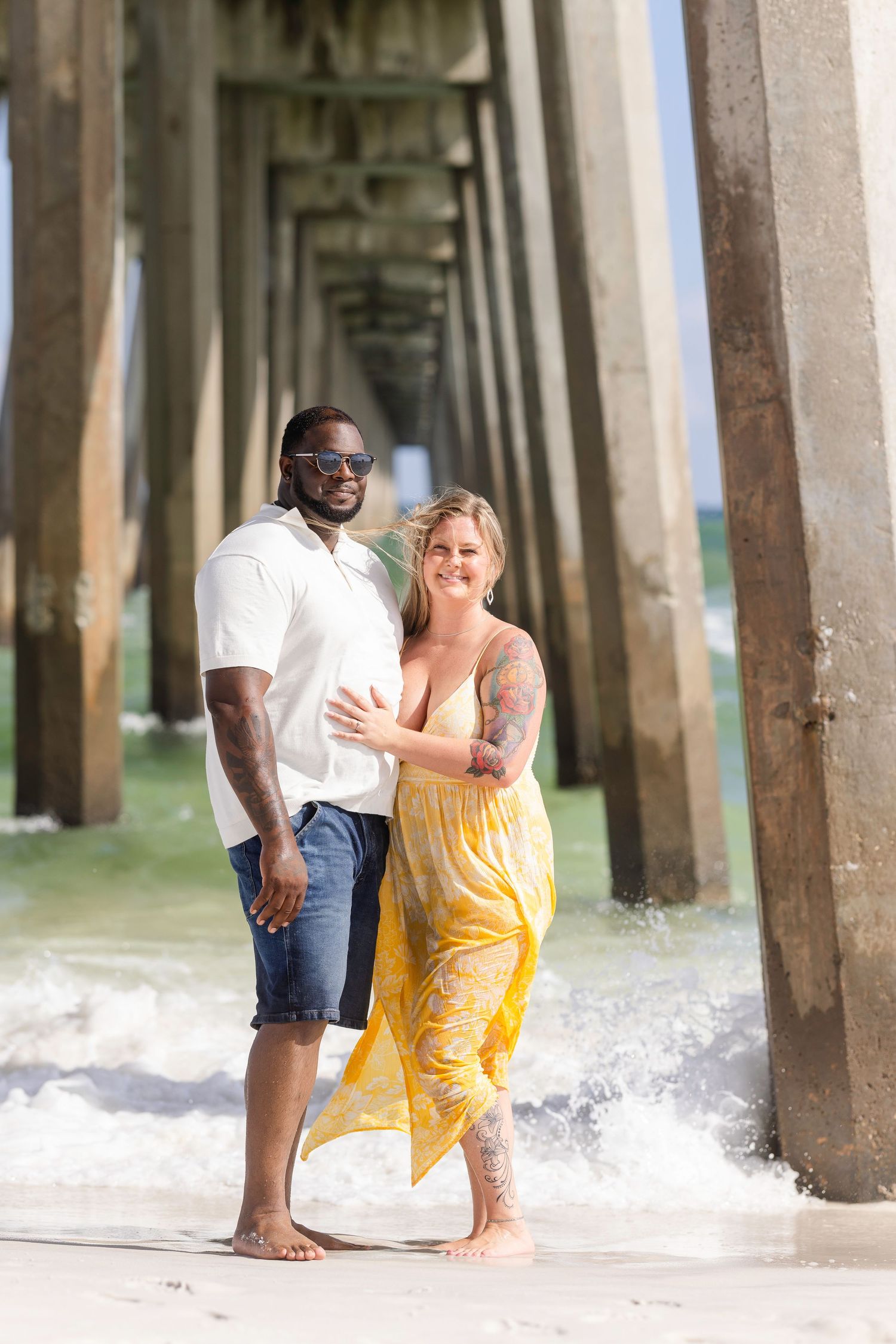 A couple embraces barefoot under a beach pier, woman in yellow dress with tattoos, man in white shirt and jeans.