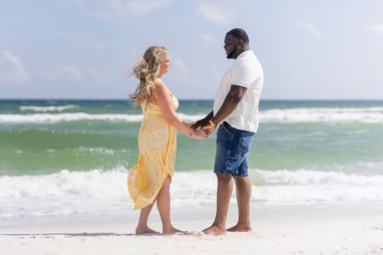 Interracial couple holding hands barefoot on a sandy beach with turquoise ocean waves in the background.