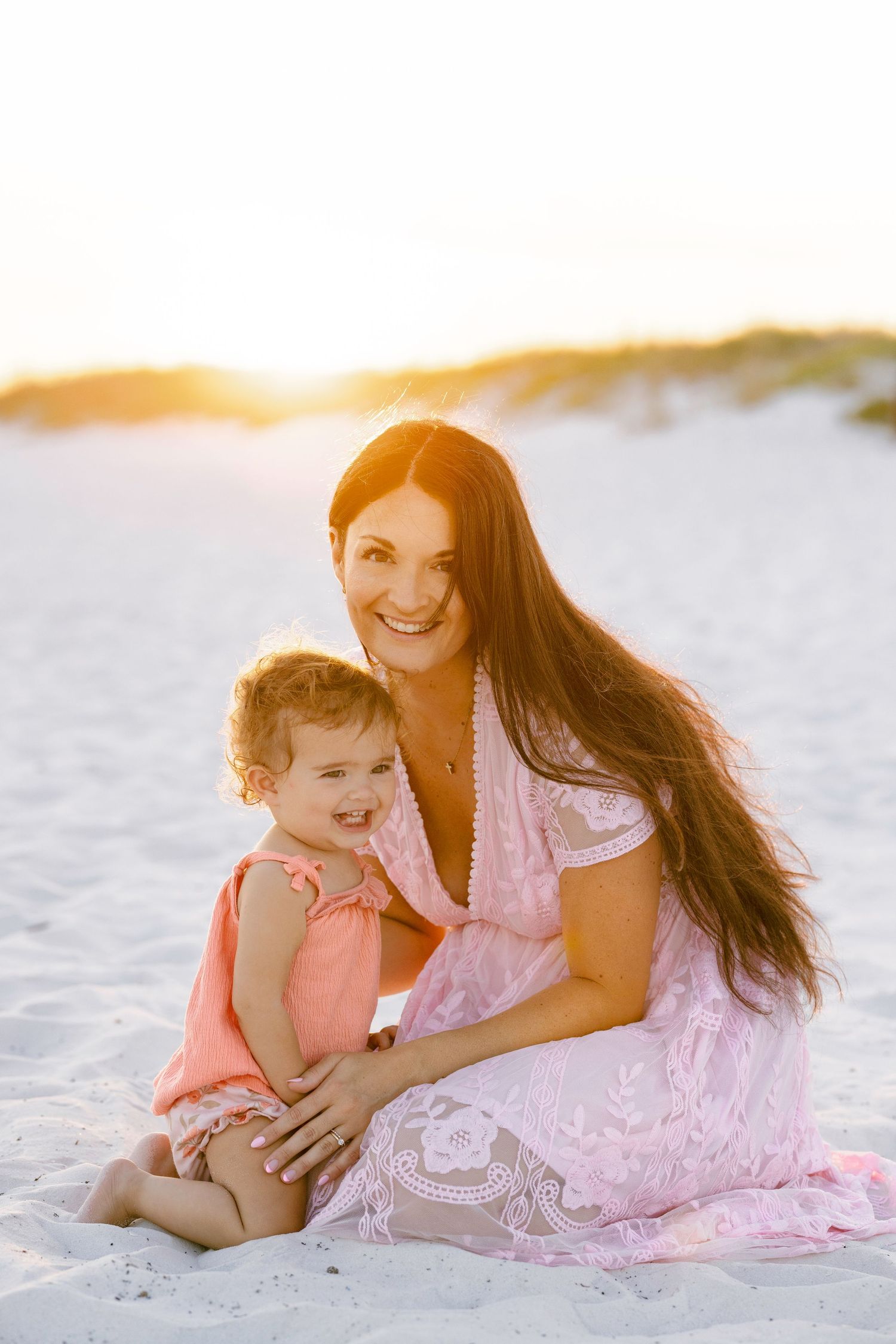 Mother in pink lace dress smiling with toddler on sandy beach at golden hour.