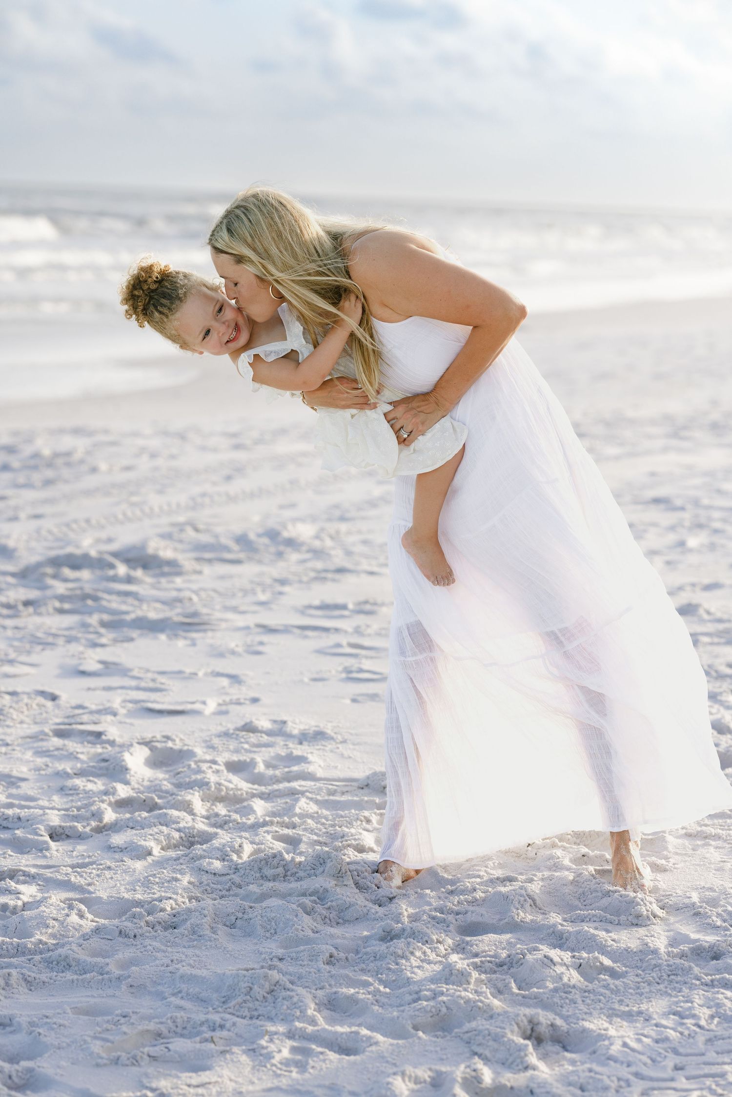 Joyful mother in white dress playing with child on sandy beach, child hugging her from behind.