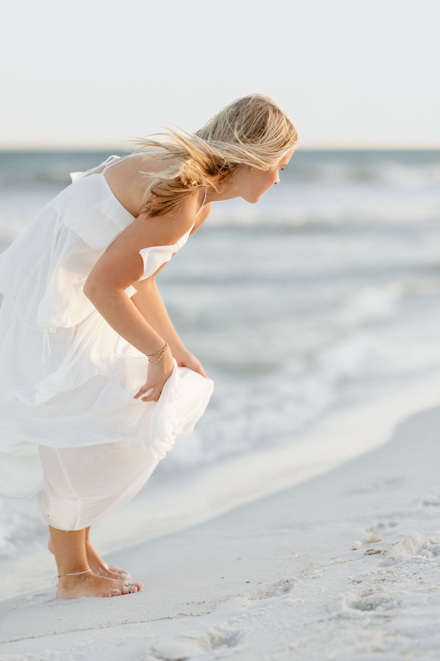 Barefoot blonde woman in flowing white dress bending down at ocean shoreline on a white sand beach.