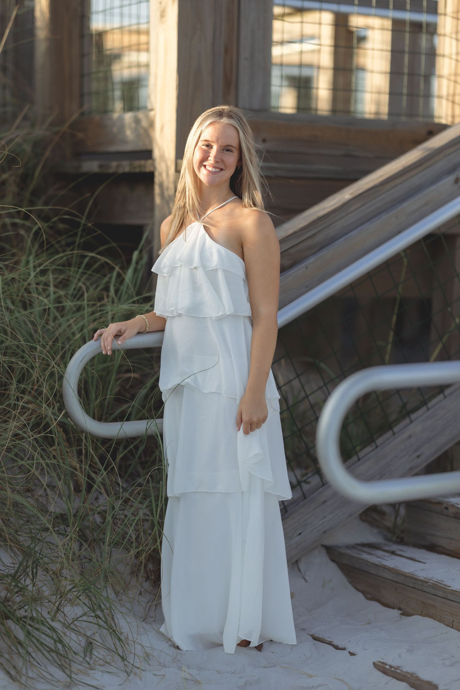 Smiling blonde woman in white tiered maxi dress posing by beach boardwalk railing with sea grass.