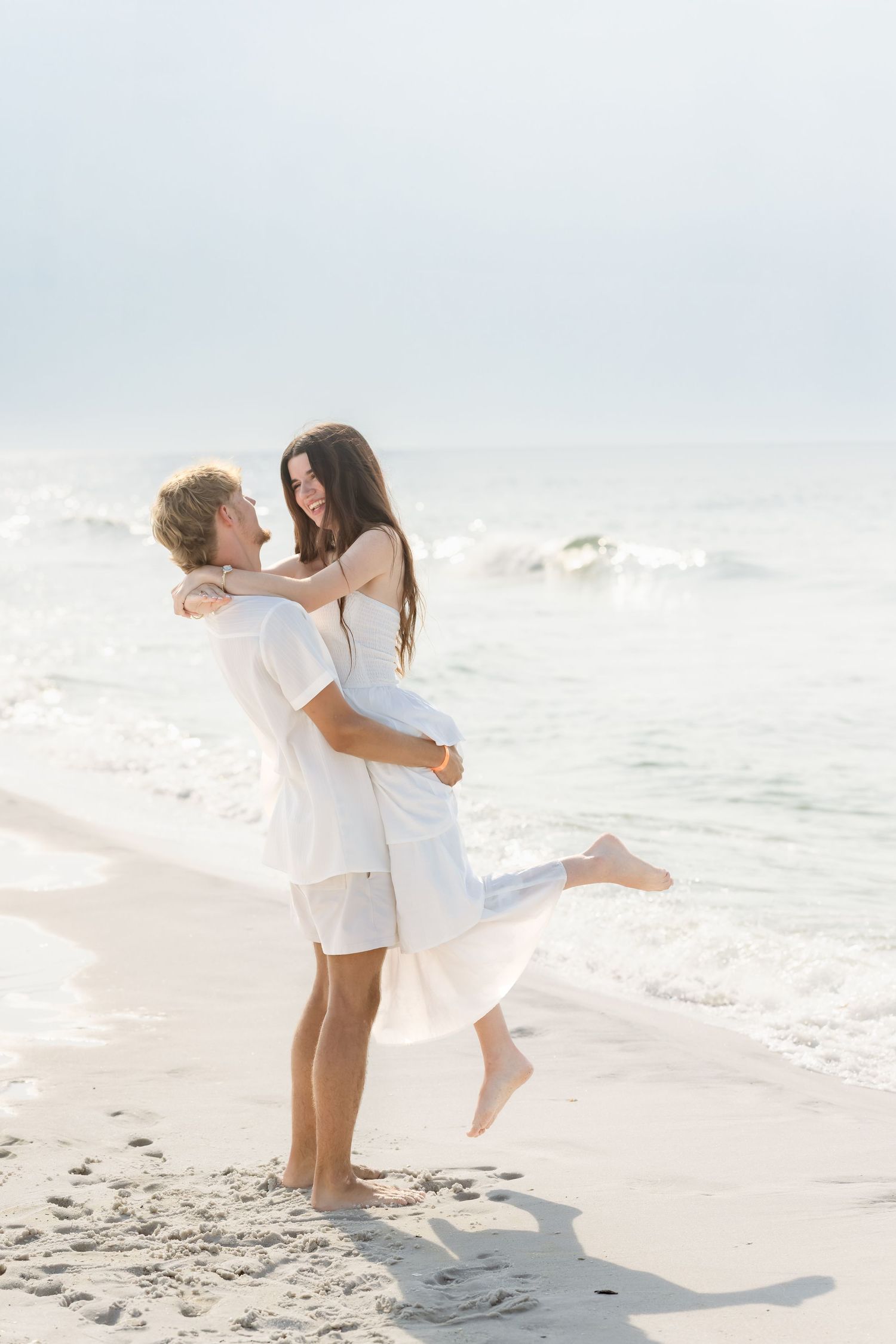 Man lifting woman in white dress on sunny beach shoreline with ocean waves behind them.