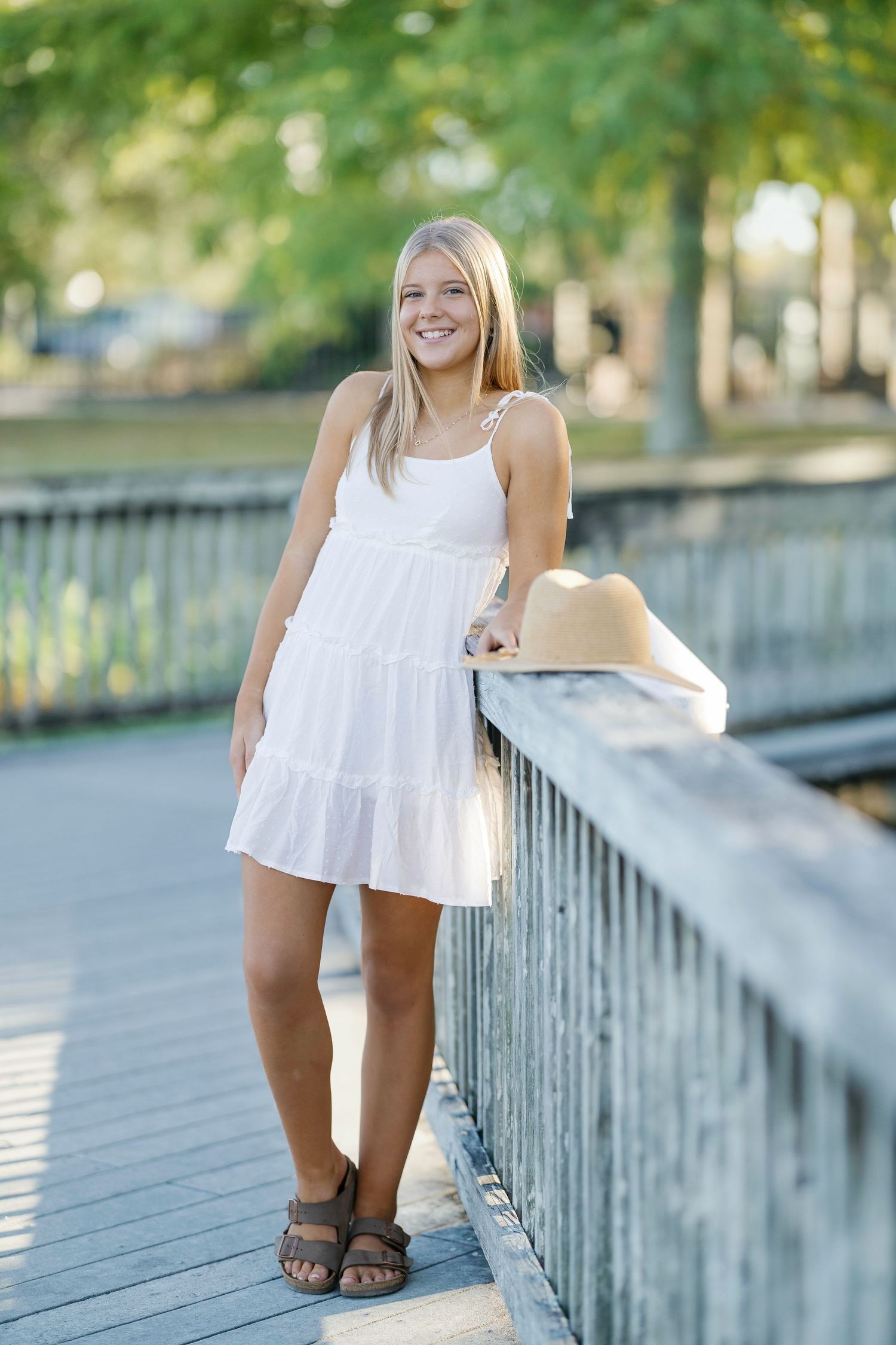Smiling young woman in white sundress holding straw hat on wooden bridge in sunny park.