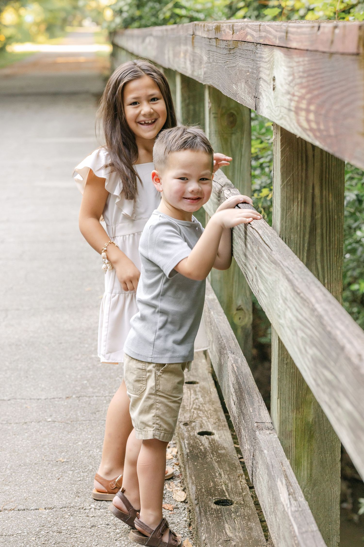 Two happy children pose by a wooden fence on a garden path, a smiling girl in white dress and boy in gray shirt.