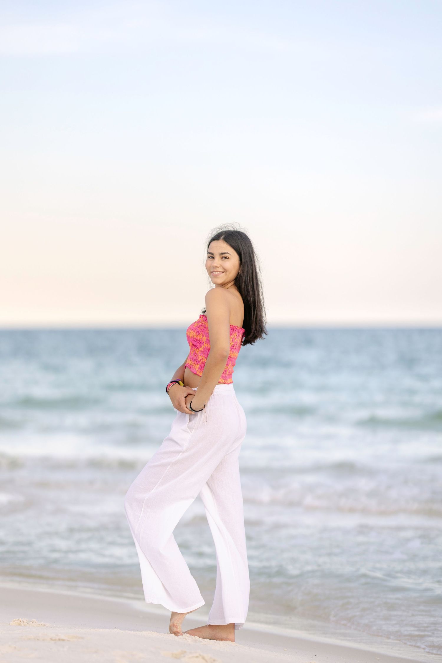 A beachside portrait showing white flowy pants and coral top against ocean waves at sunset.