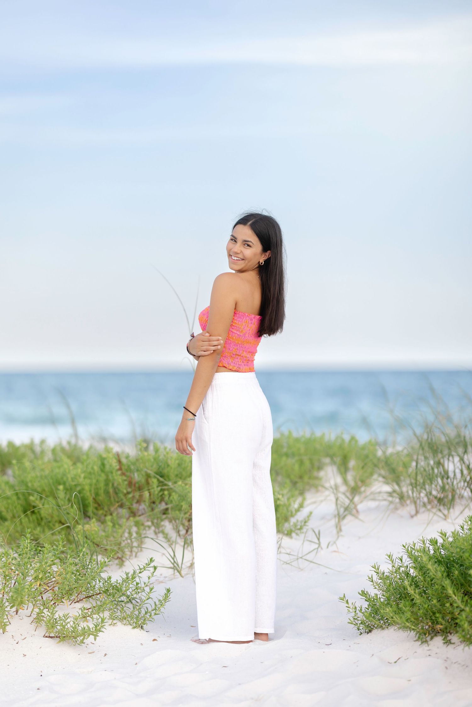 Woman wearing white skirt and coral top poses on beach dunes near ocean.