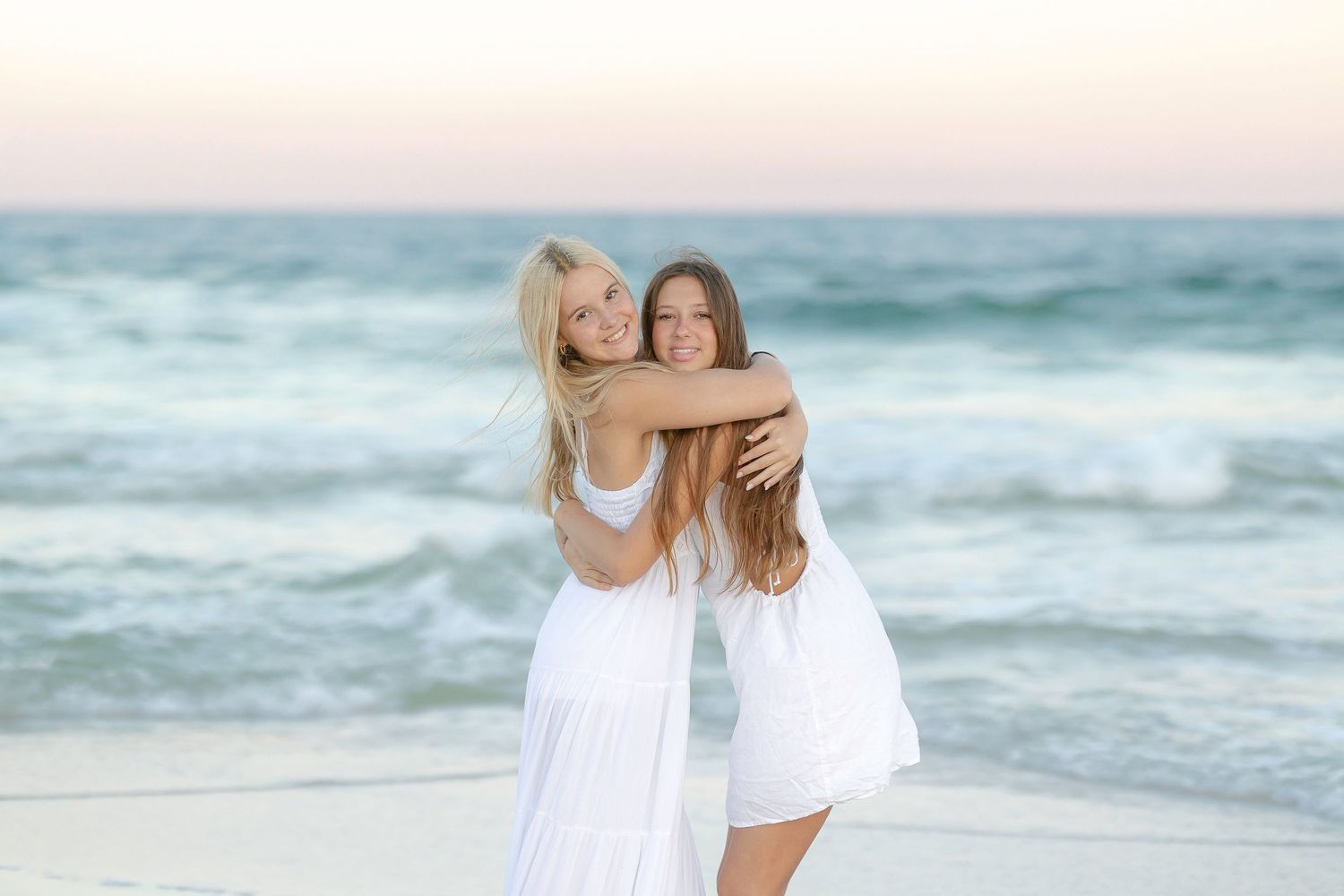 Friends share a warm hug while wearing matching white dresses on a beach at dusk.