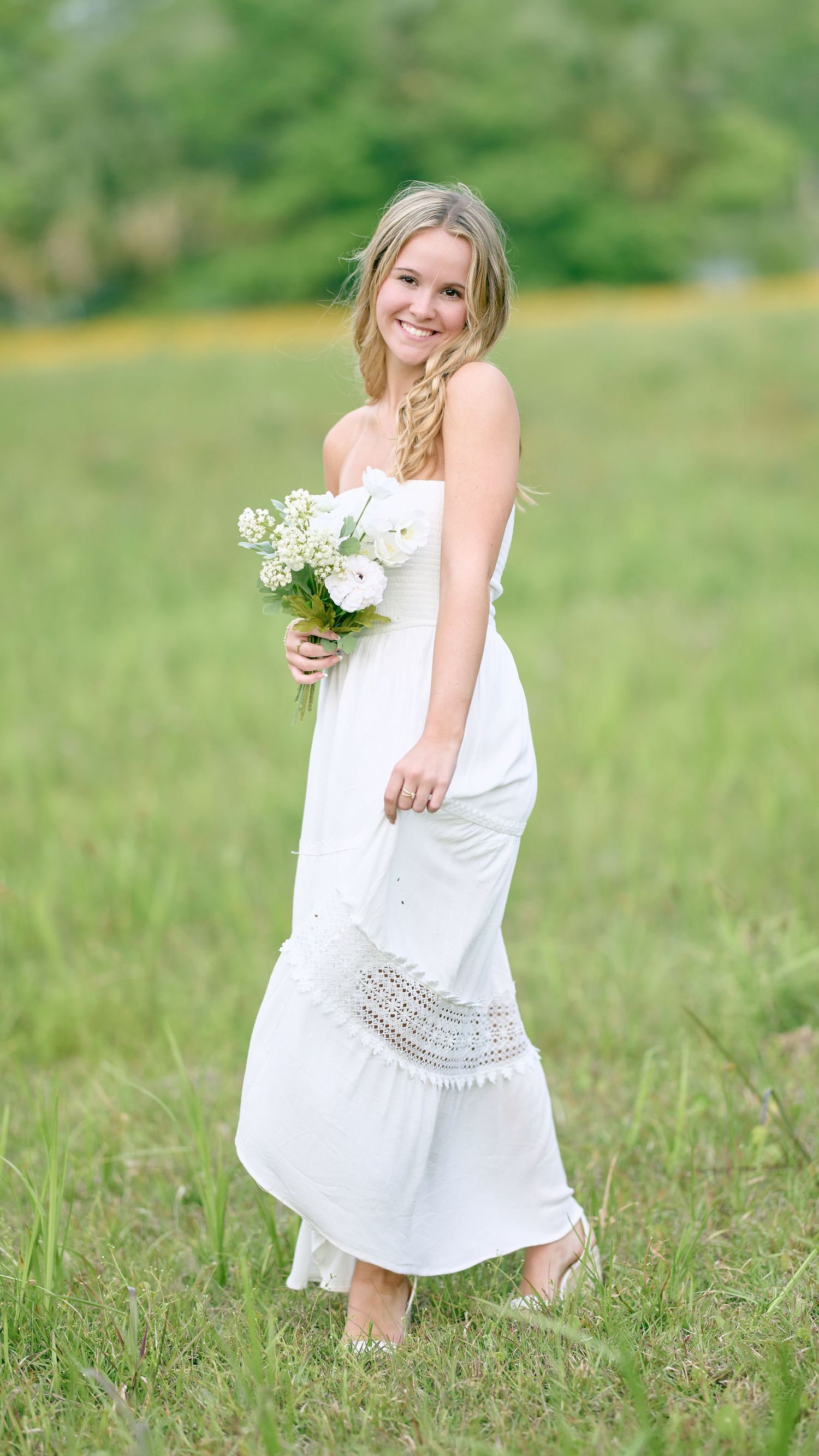 Woman in white maxi dress holding bouquet while standing in green field.