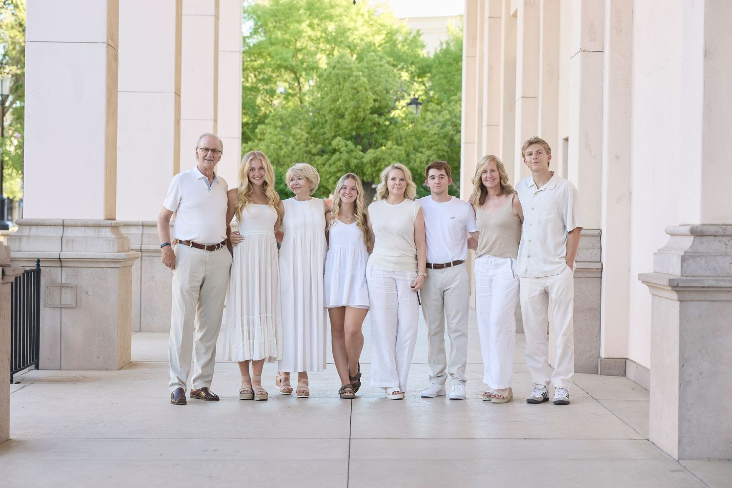 A group in coordinated white and beige outfits poses together between elegant stone columns on a sunny day.