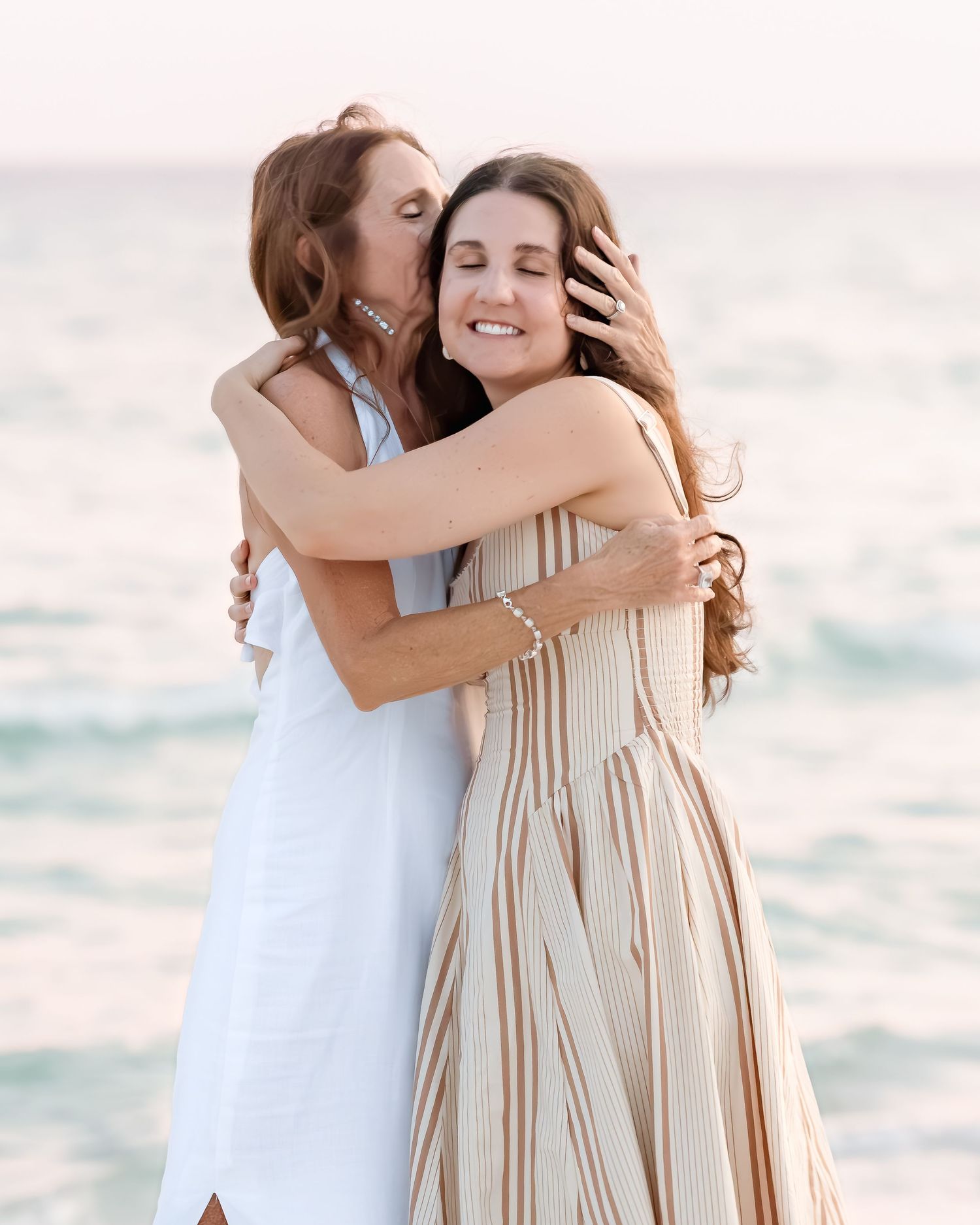 Two women embracing in a warm hug on a beach, one in white dress, other in striped sundress, ocean backdrop.