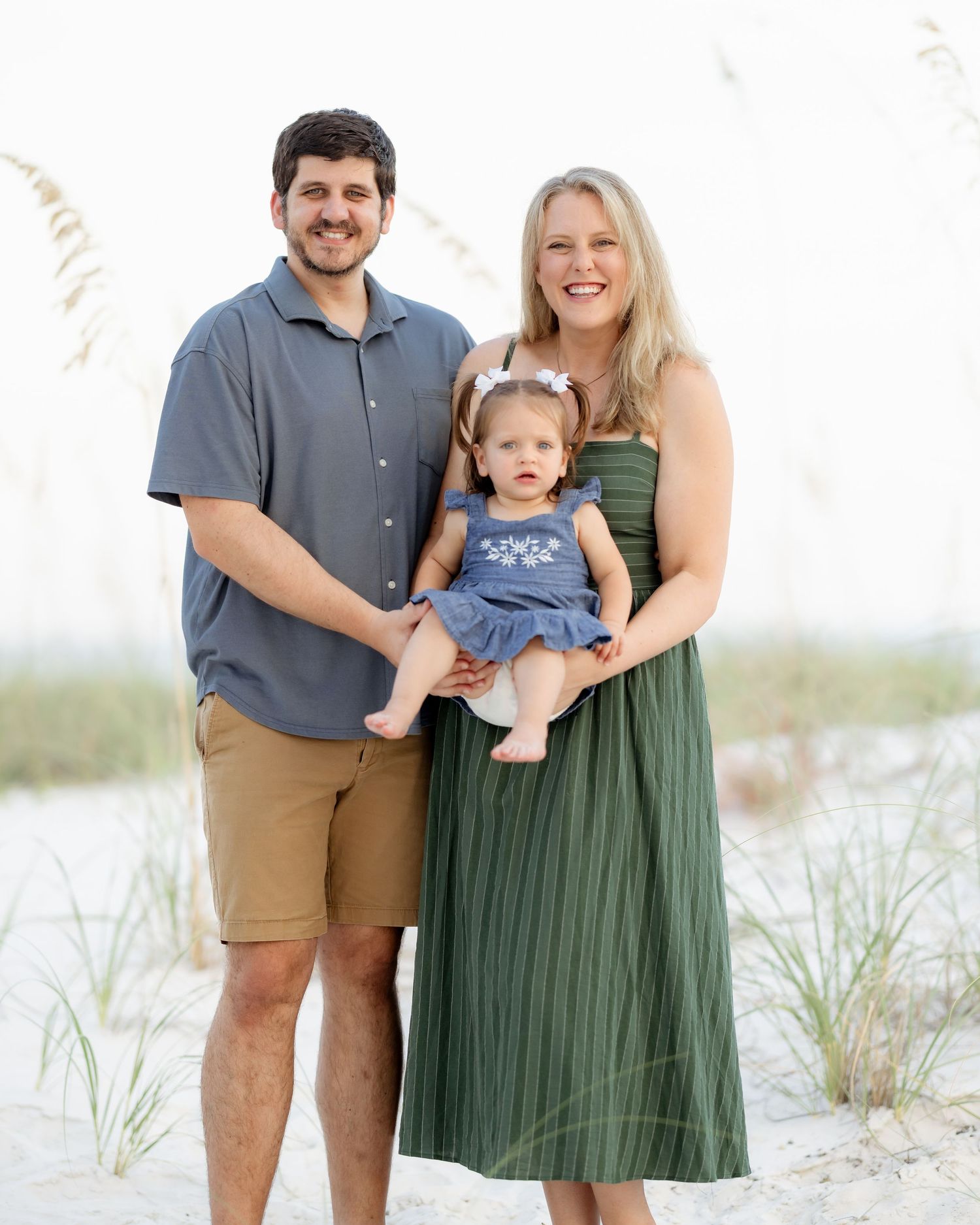 A smiling family of three poses on a sandy beach, with parents holding their baby girl in a blue dress.
