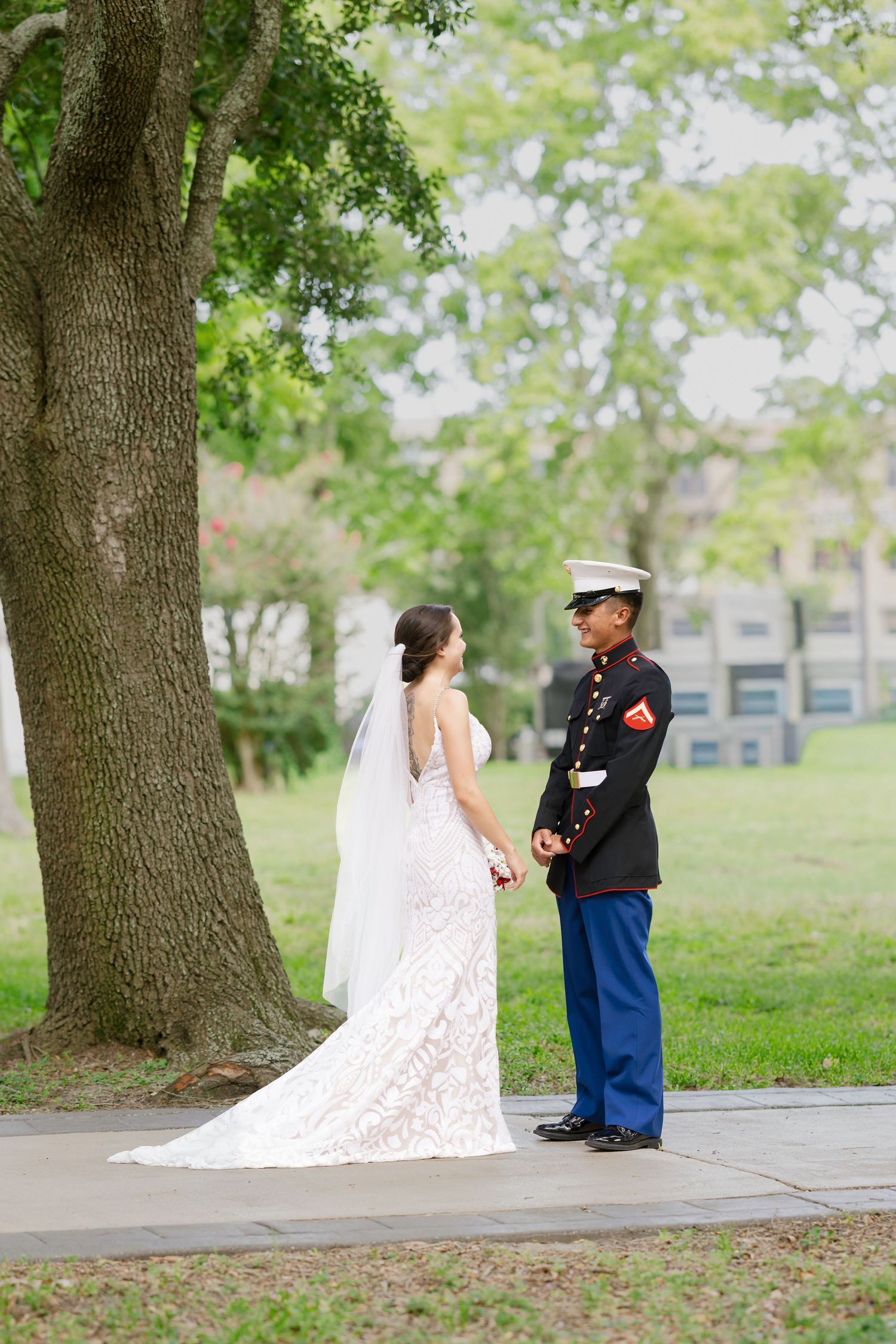 A bride in a lace gown and Marine groom in dress blues share a first look outdoors near a large tree.