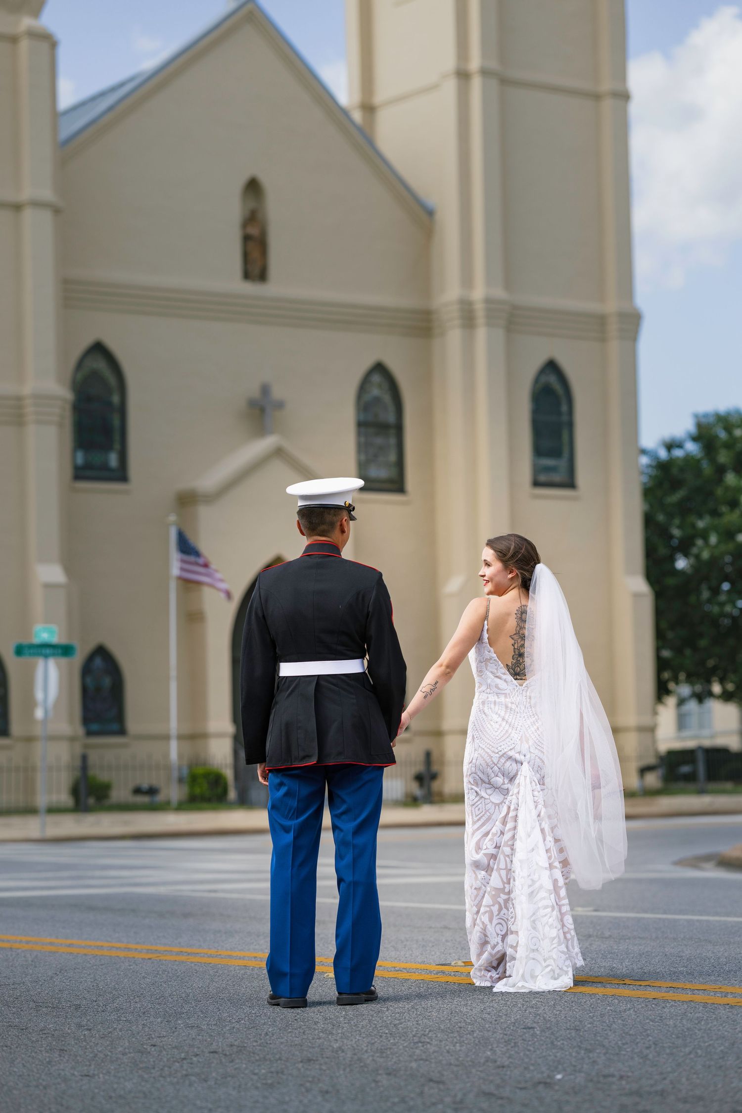 Marine groom in dress blues holds bride's hand in white lace gown outside church on wedding day.