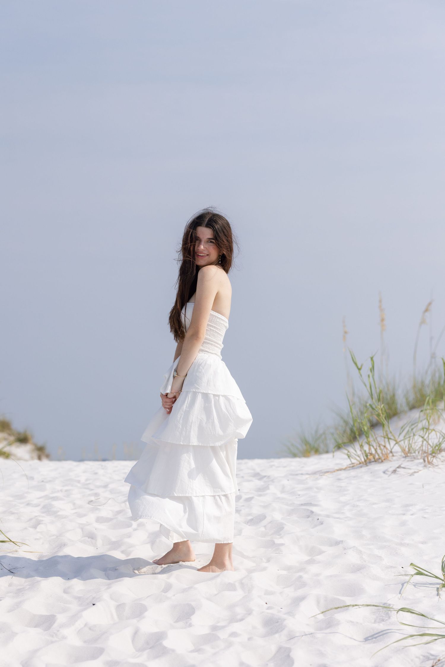 Young woman in white strapless tiered maxi dress standing barefoot on white sand dunes with sea grass.